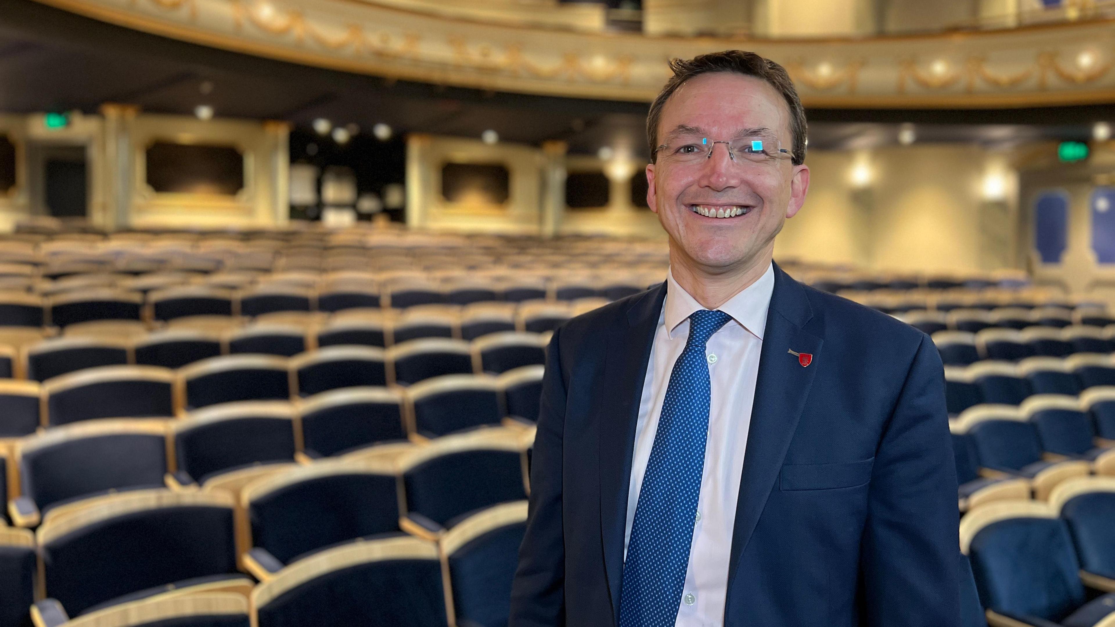 A man wearing a navy suit white shirt and light blue tie, standing in a theatre with rows of chairs behind him.