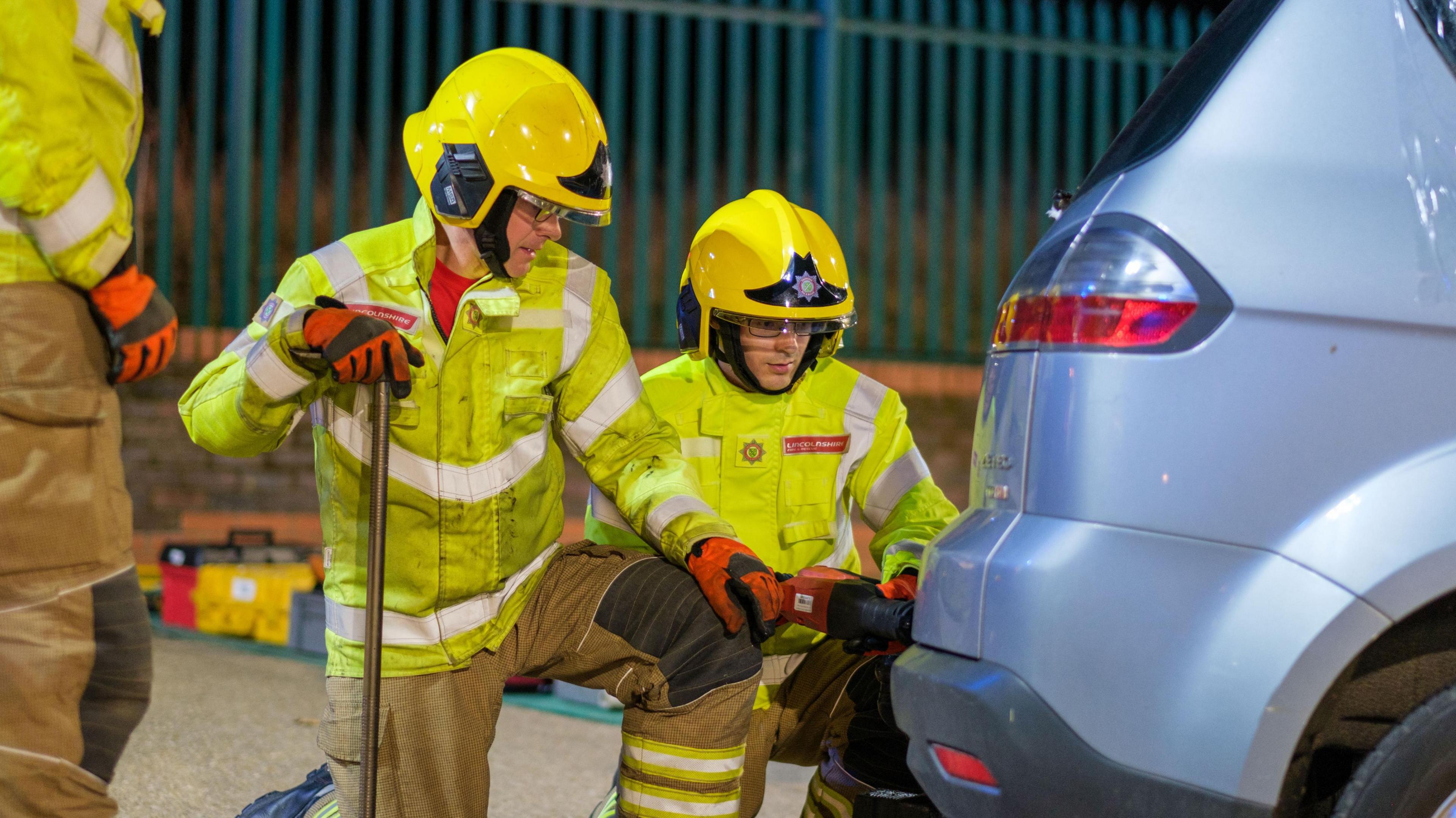 Two firefighters wearing yellow hi-visibility jackets, yellow hard hats and beige trousers knelt on the floor outside working to open the boot of a silver car with tools. 