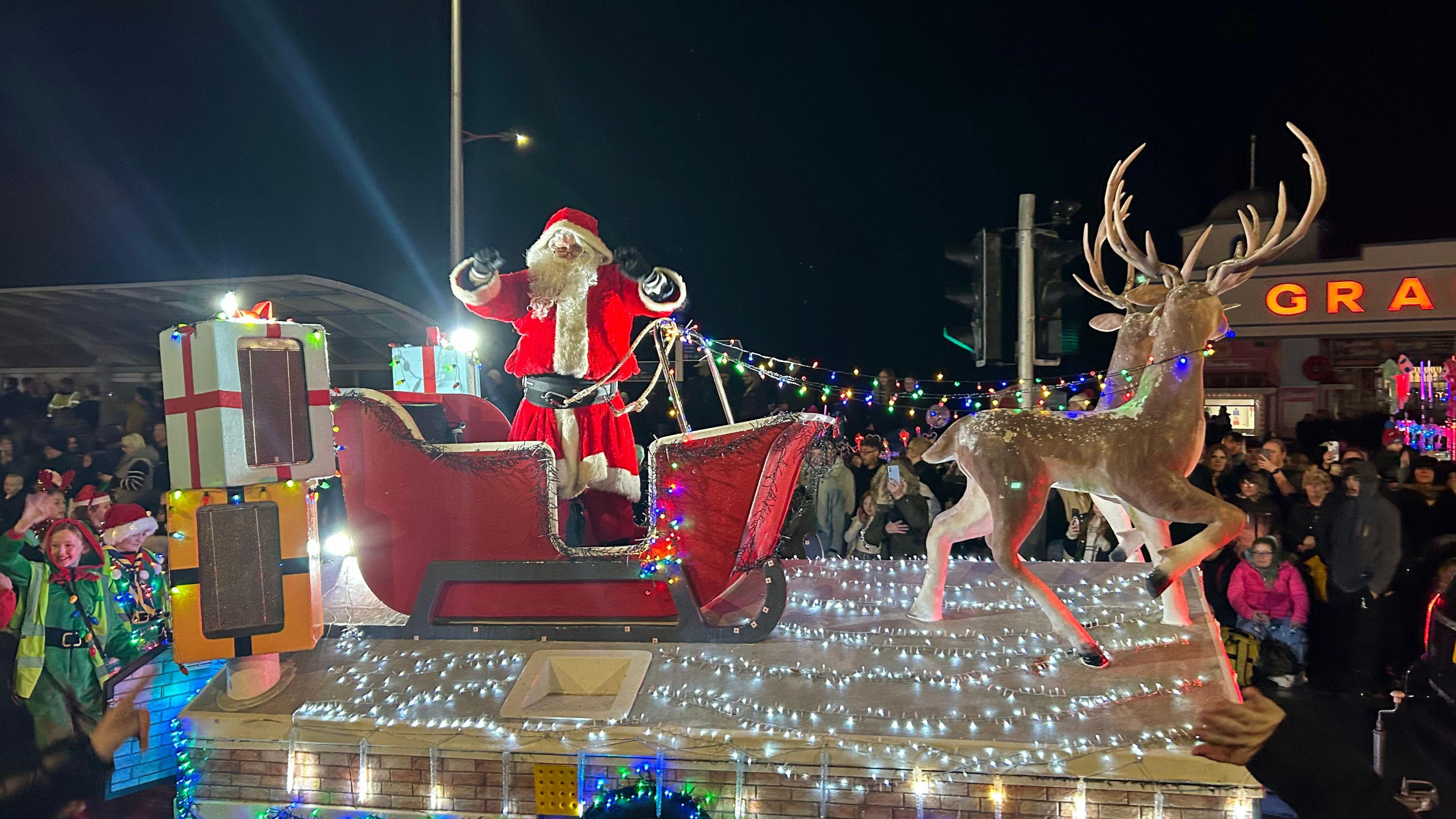 A Christmas-themed carnival cart. Somebody dressed as Santa is sat in a sleigh behind two model reindeers.