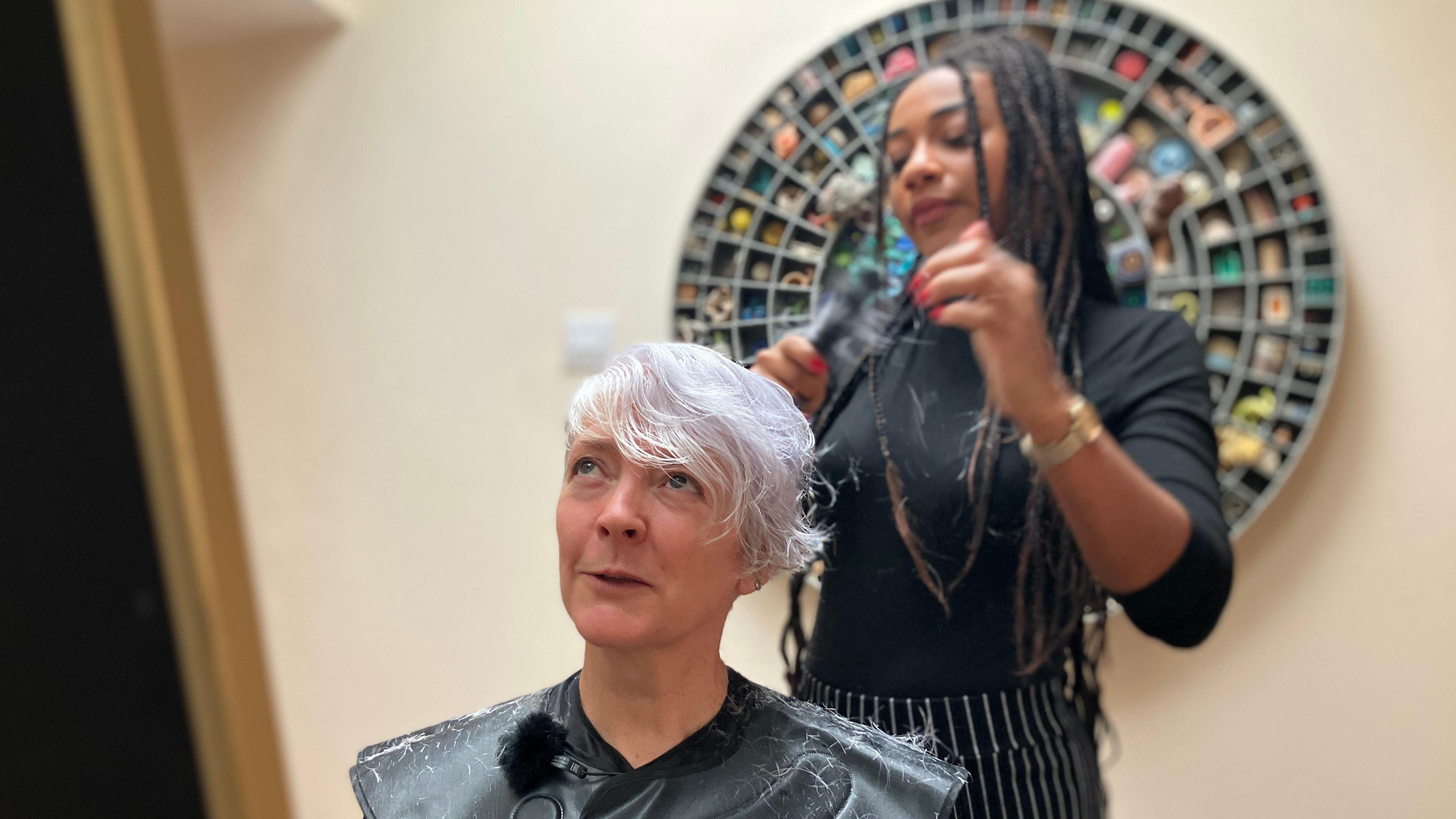 A woman sat down, with short hair, wearing a cape and having her hair cut by a woman stood behind her wearing all black. 