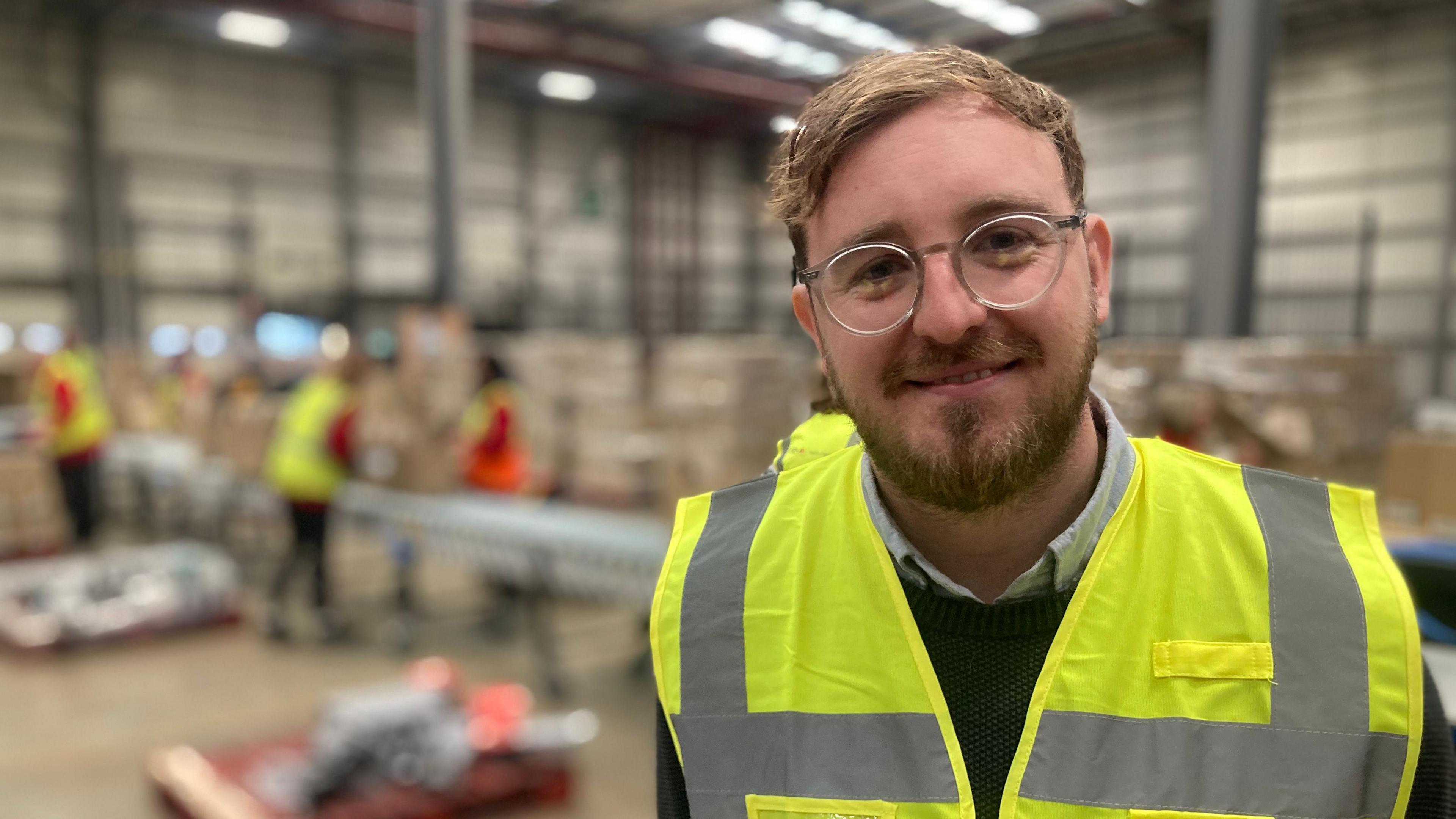 A man with dark blonde swept back hair and a beard wearing a high visibility jacket stands in a warehouse with people packing in the background