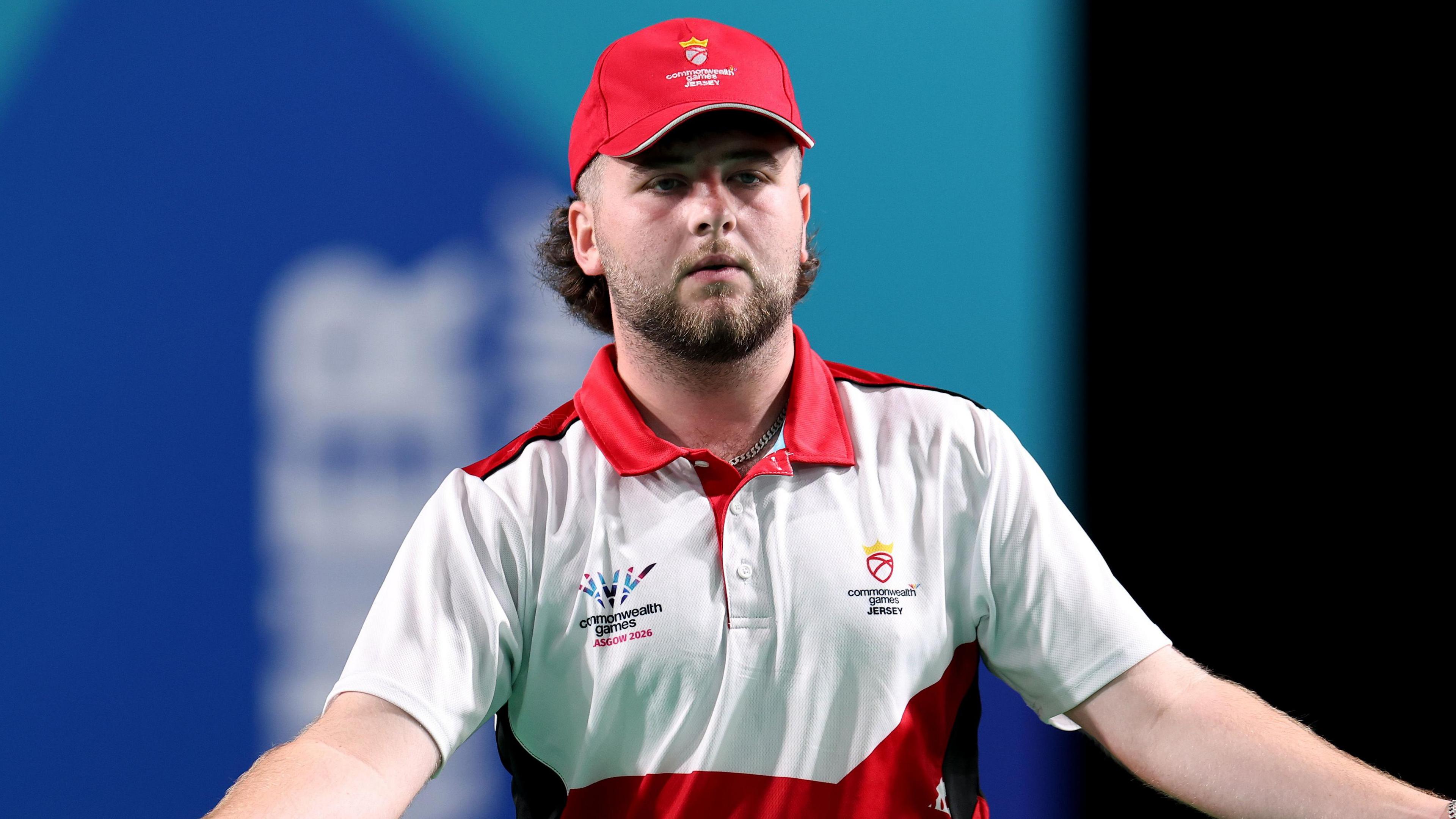 Taylor Greechan wearing a Jersey Commonwealth Games branded white and red polo top and red cap stands with his arms outstretched