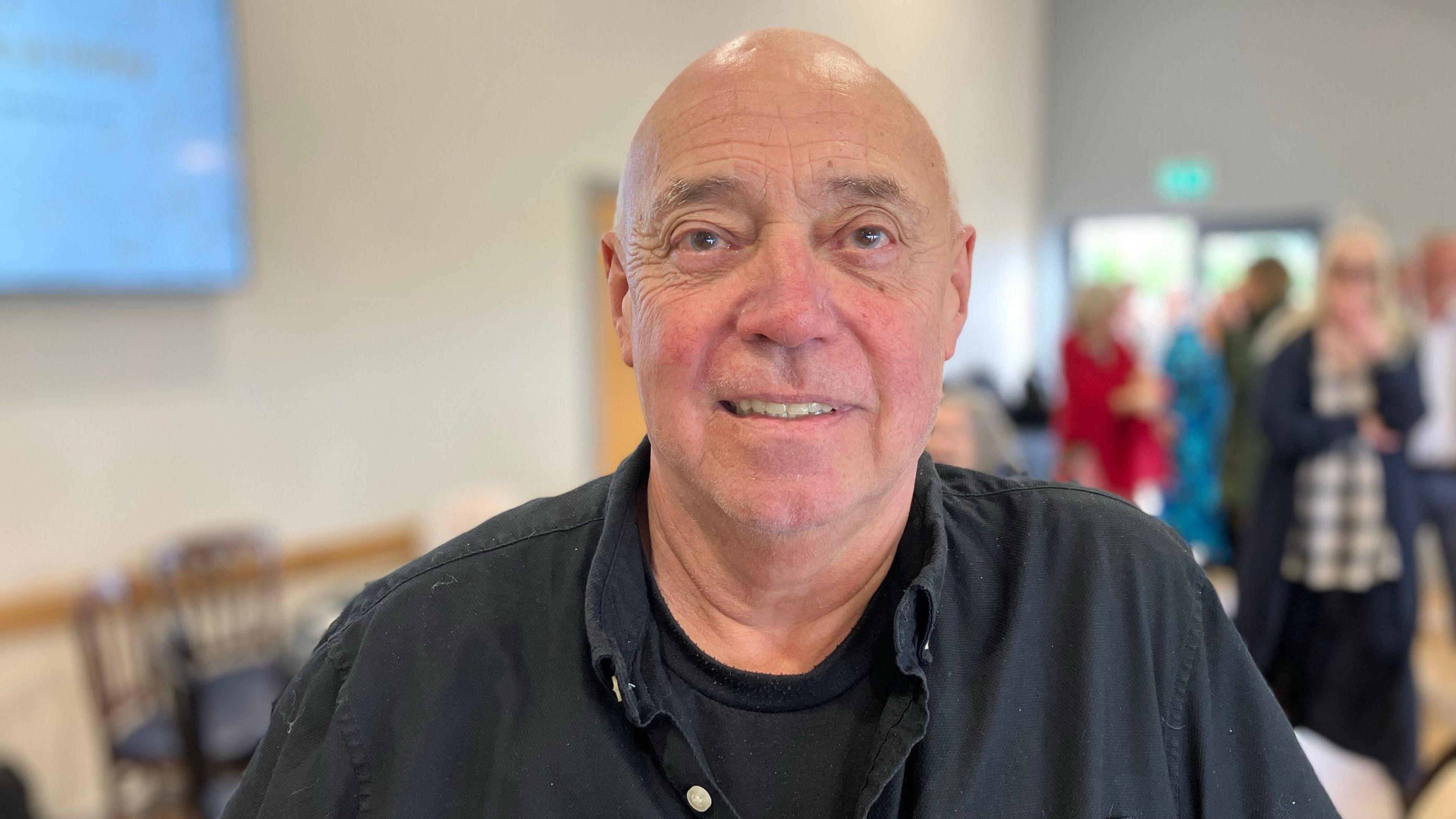 Alan Hudson is bald, and is smiling at the camera. He is wearing a balack button-up t-shirt