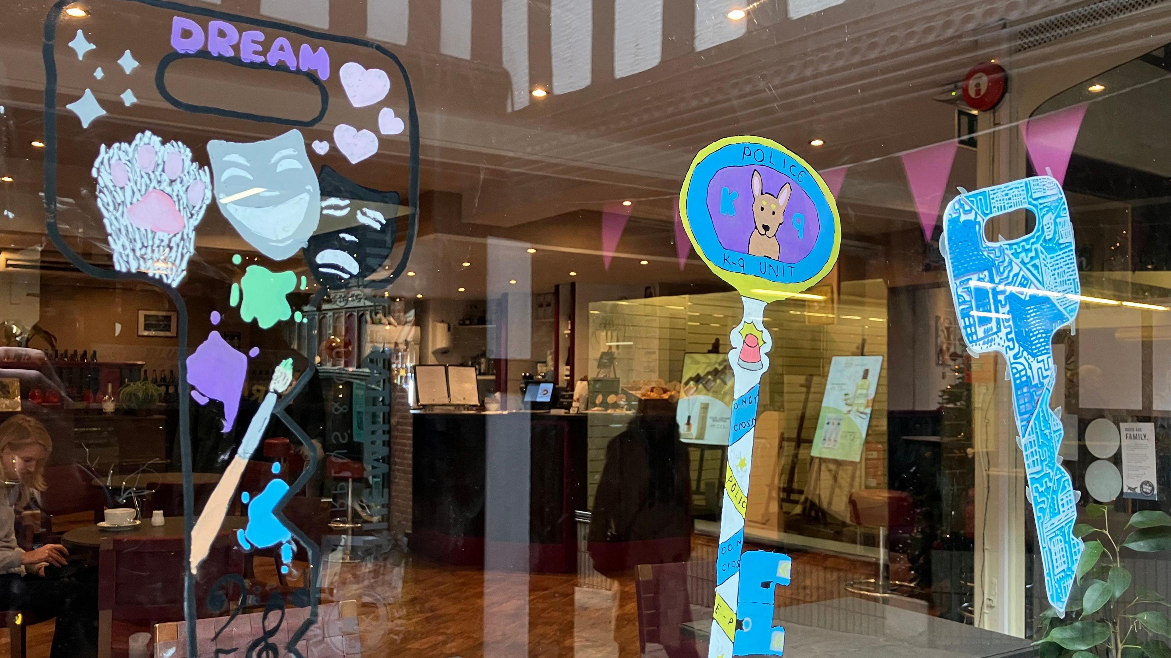 Three key-shaped pictures in a cafe window, one has a police dog, another has the word Dream and a paint brush.