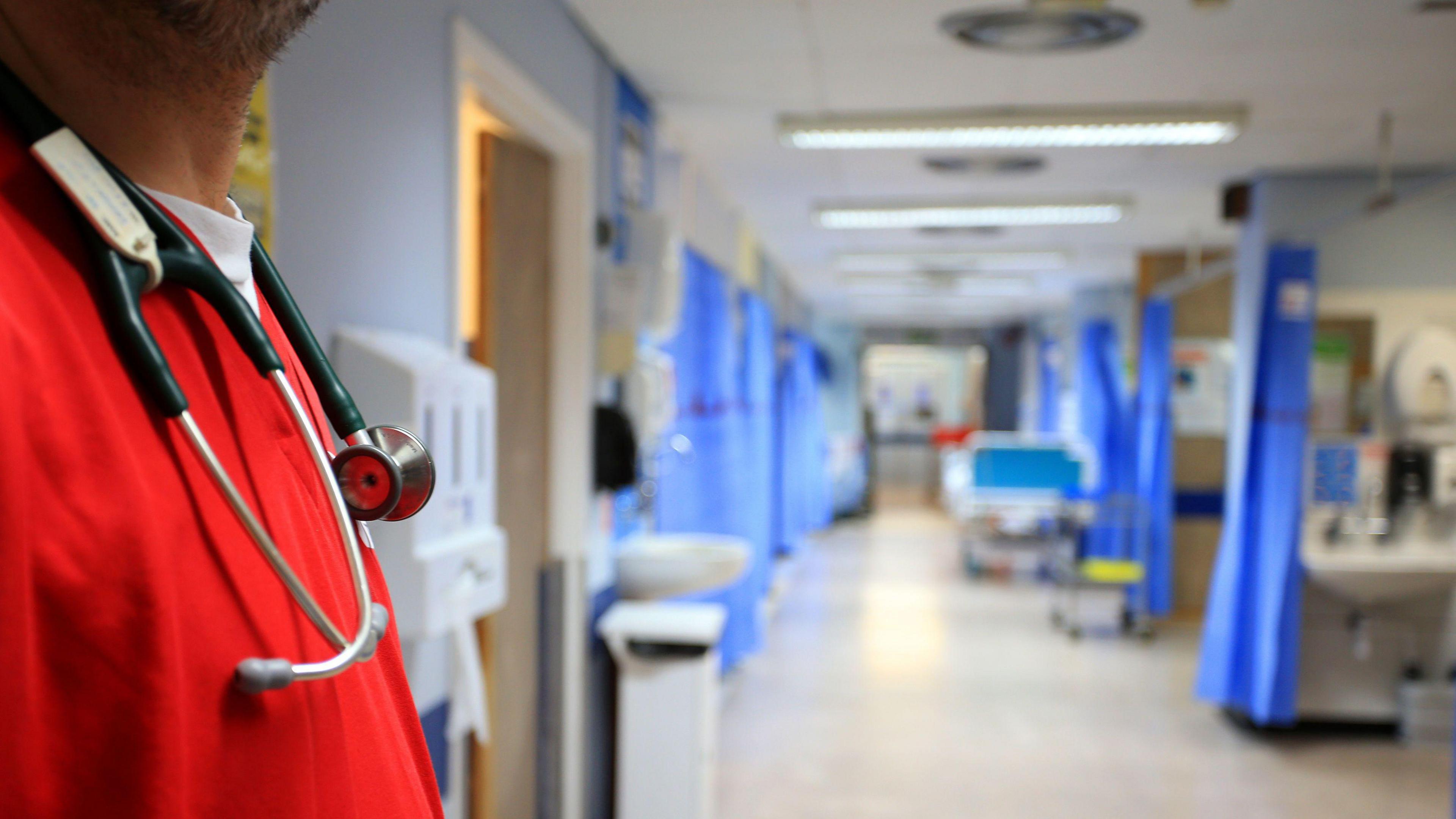 A hospital corridor, with booths either side screened by blue curtains. In the foreground is a male doctor wearing red scrubs and a stethoscope around his neck. 