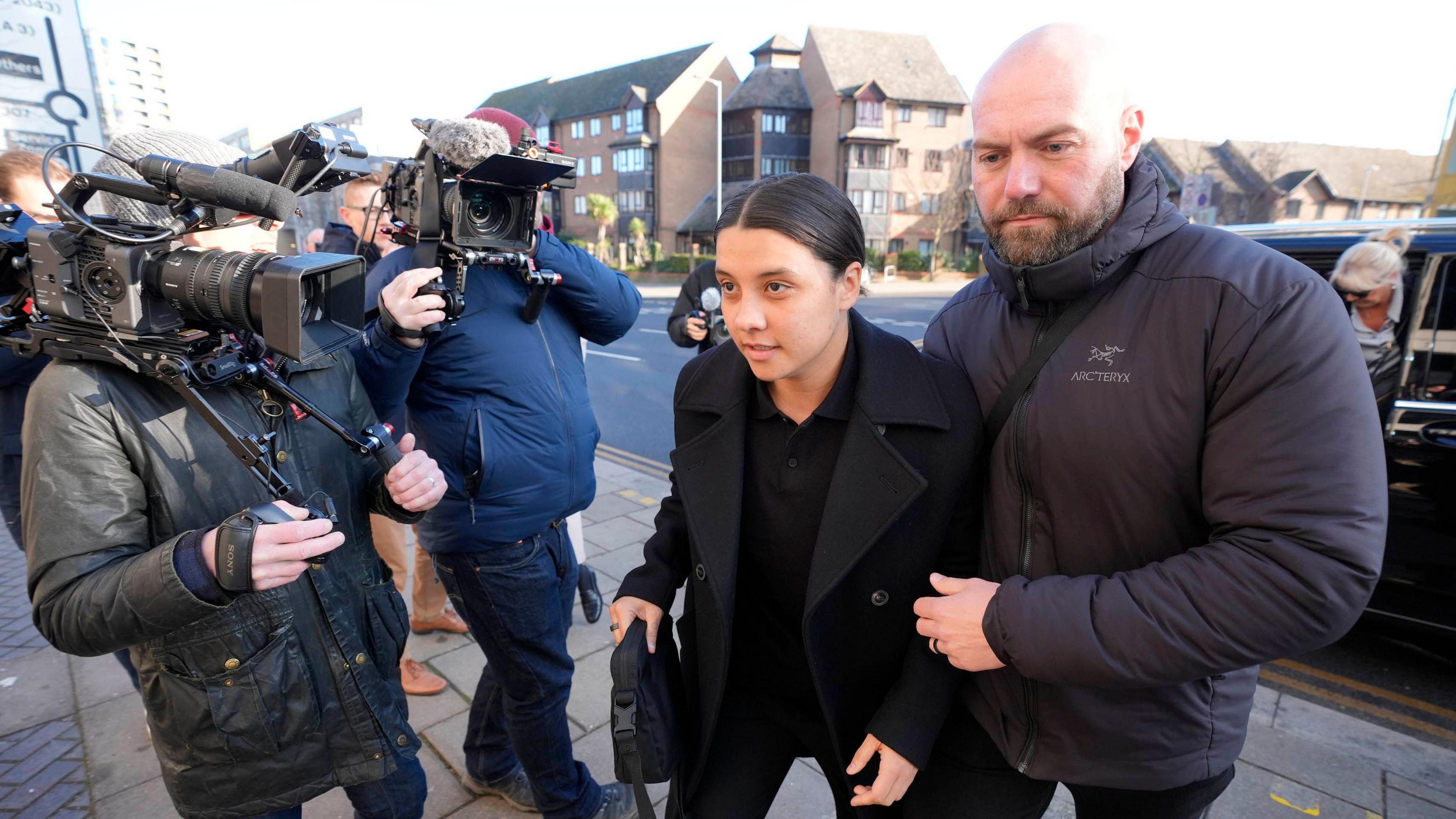 Chelsea and Australia striker Sam Kerr at Kingston Crown Court, south-west London. TV cameras are pointed at her. She's being led by a bodyguard.