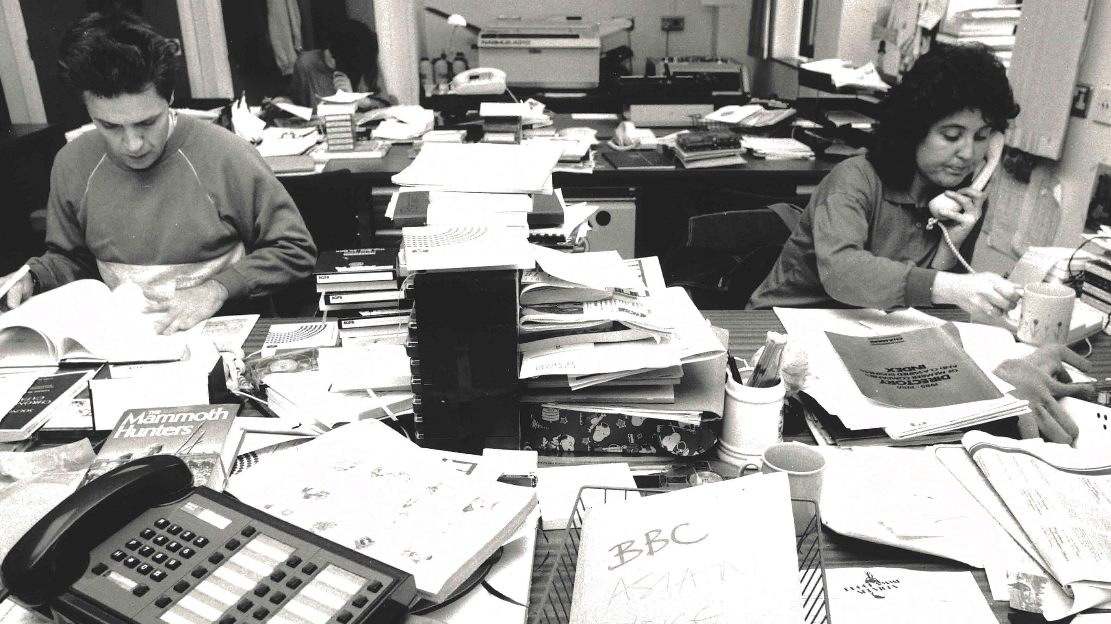 A man and a women sitting at a large desk with a huge pile of paperwork in front of them. The woman is on the phone and reaching for a cup of tea. The office is messy.