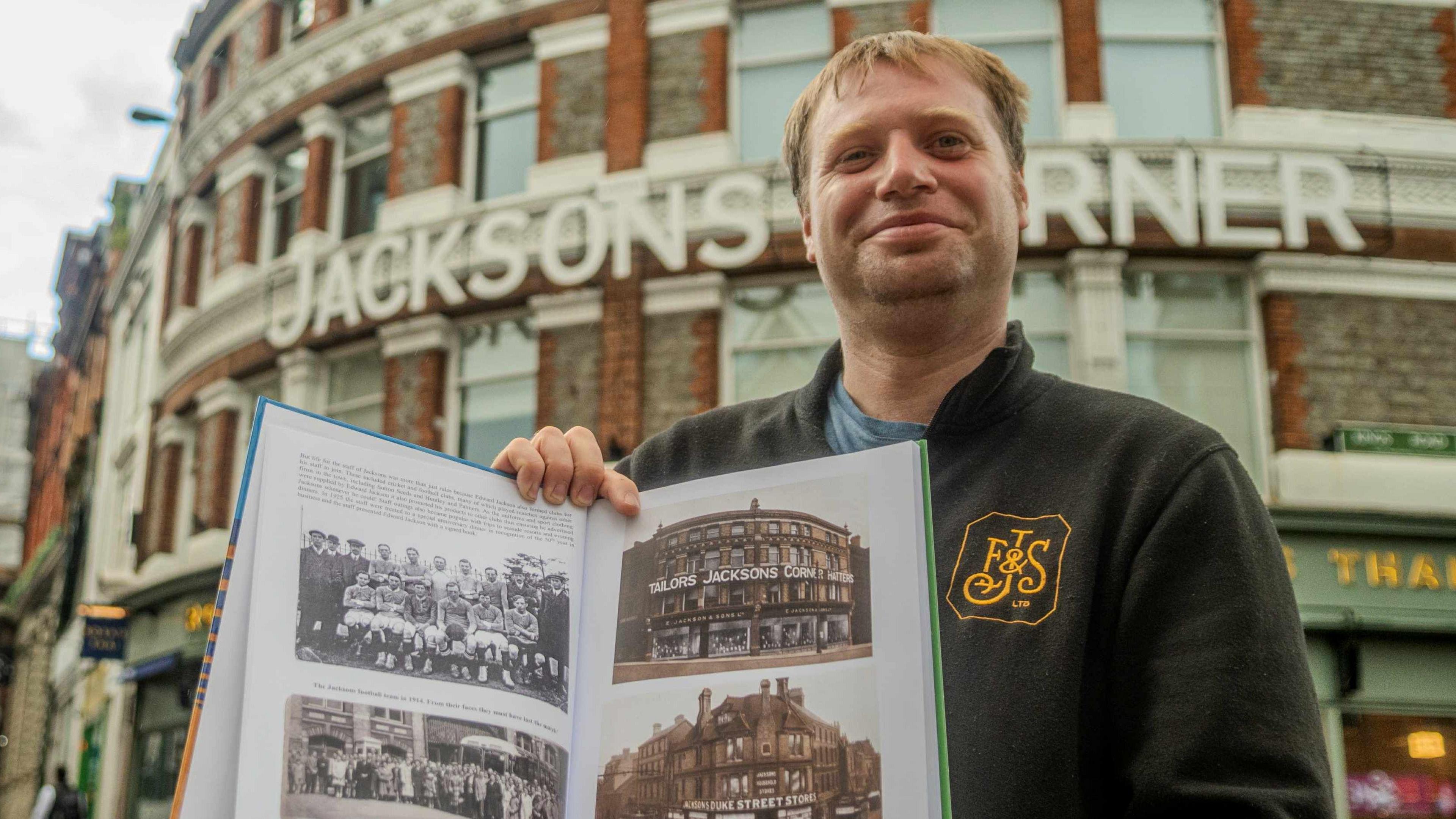 Thomas Macey stood in front of the Jacksons Corner site holding an open booklet with photographs of the old store which he is presenting to the camera.
