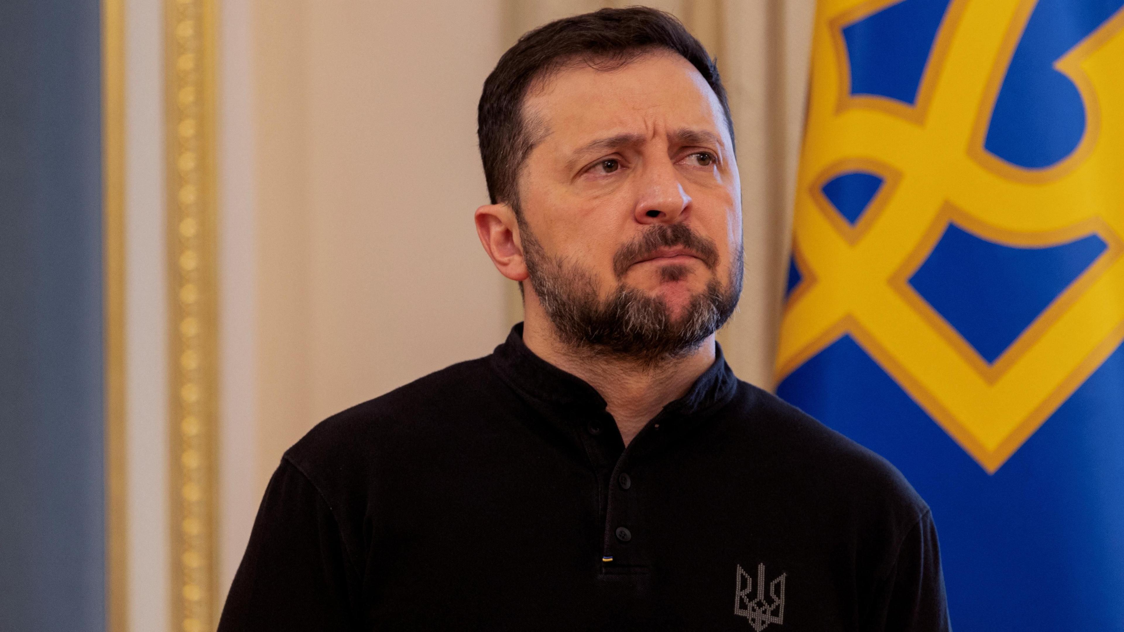 Volodymyr Zelensky, wearing a black top with a symbol embroidered on it, stands in front of a blue flag with a yellow symbol on it