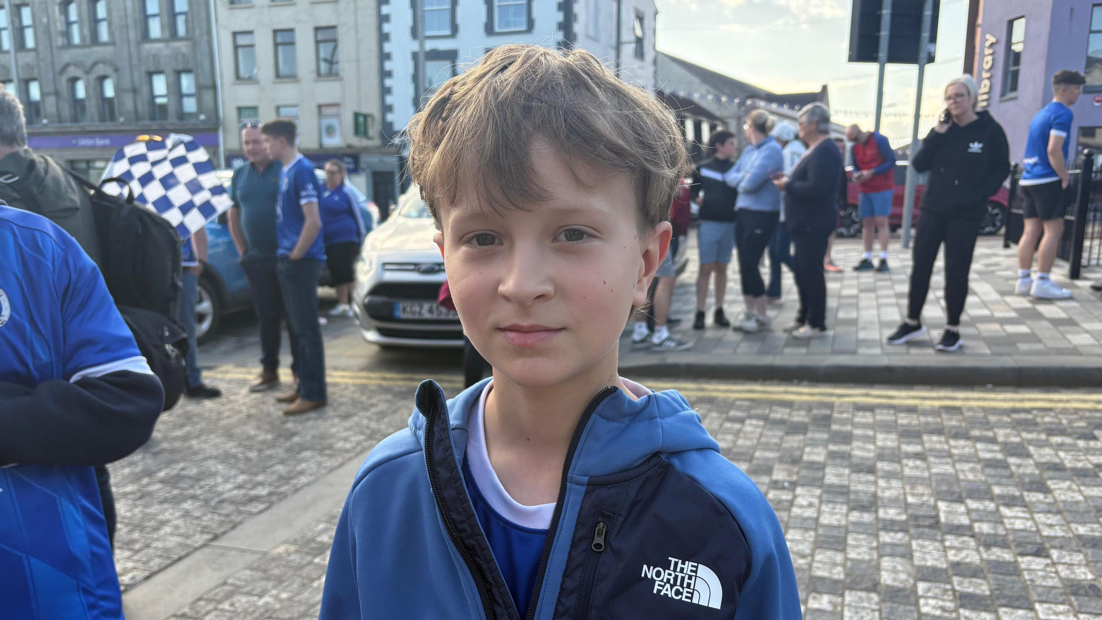 James. He has light brown hair, wearing a blue North Face jacket and blue Swifts jersey underneath. Other people are in the background.