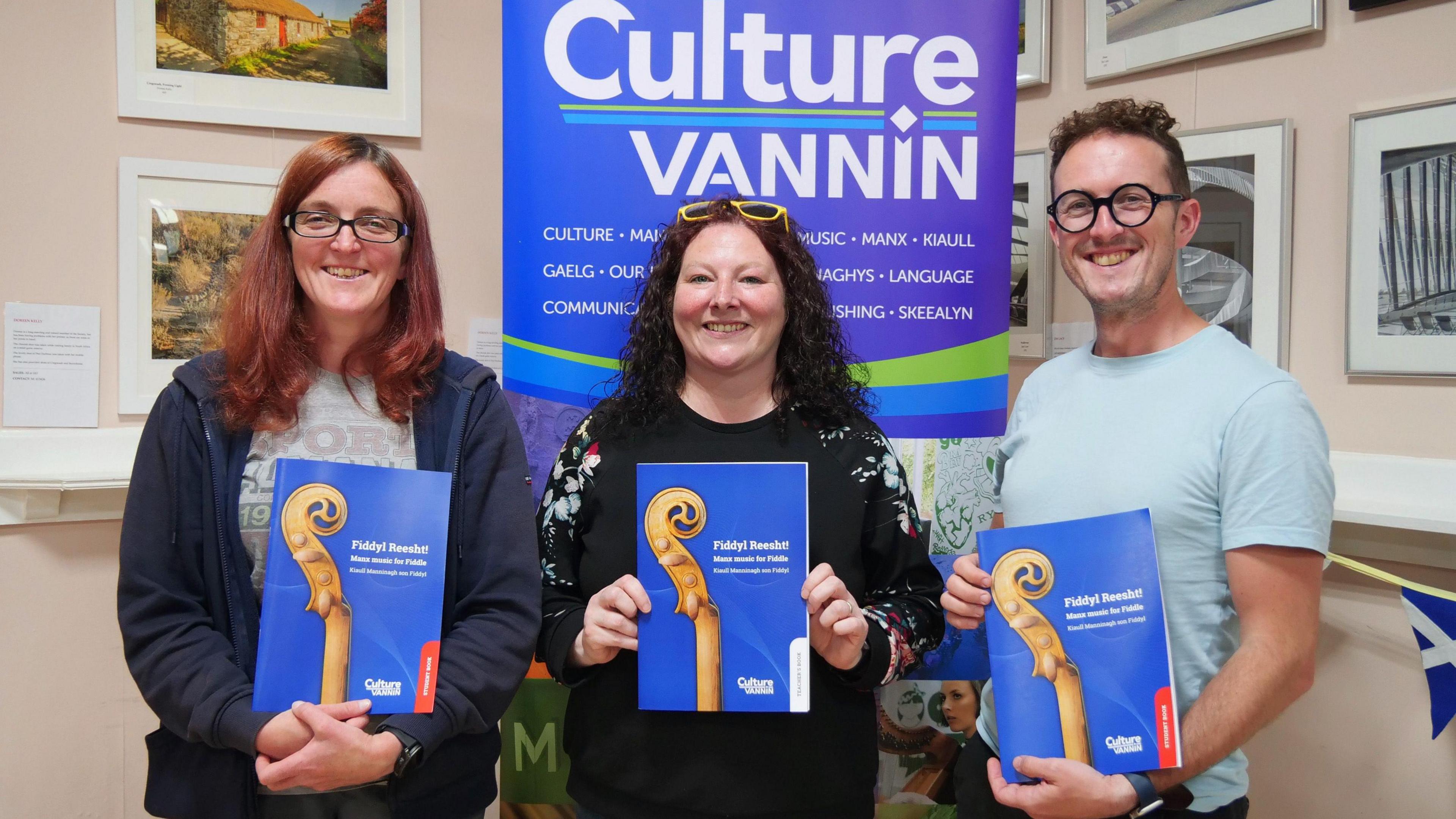 Katie Lawrence, a woman with light brown hair and glasses, Laura Rowles who has curly dark brown hair, and David Kigallon, who has short brown hair and glasses. They all smile while holding the blue book which has the head of a violin on the cover. They are standing in front of a banner that reads Culture Vannin.