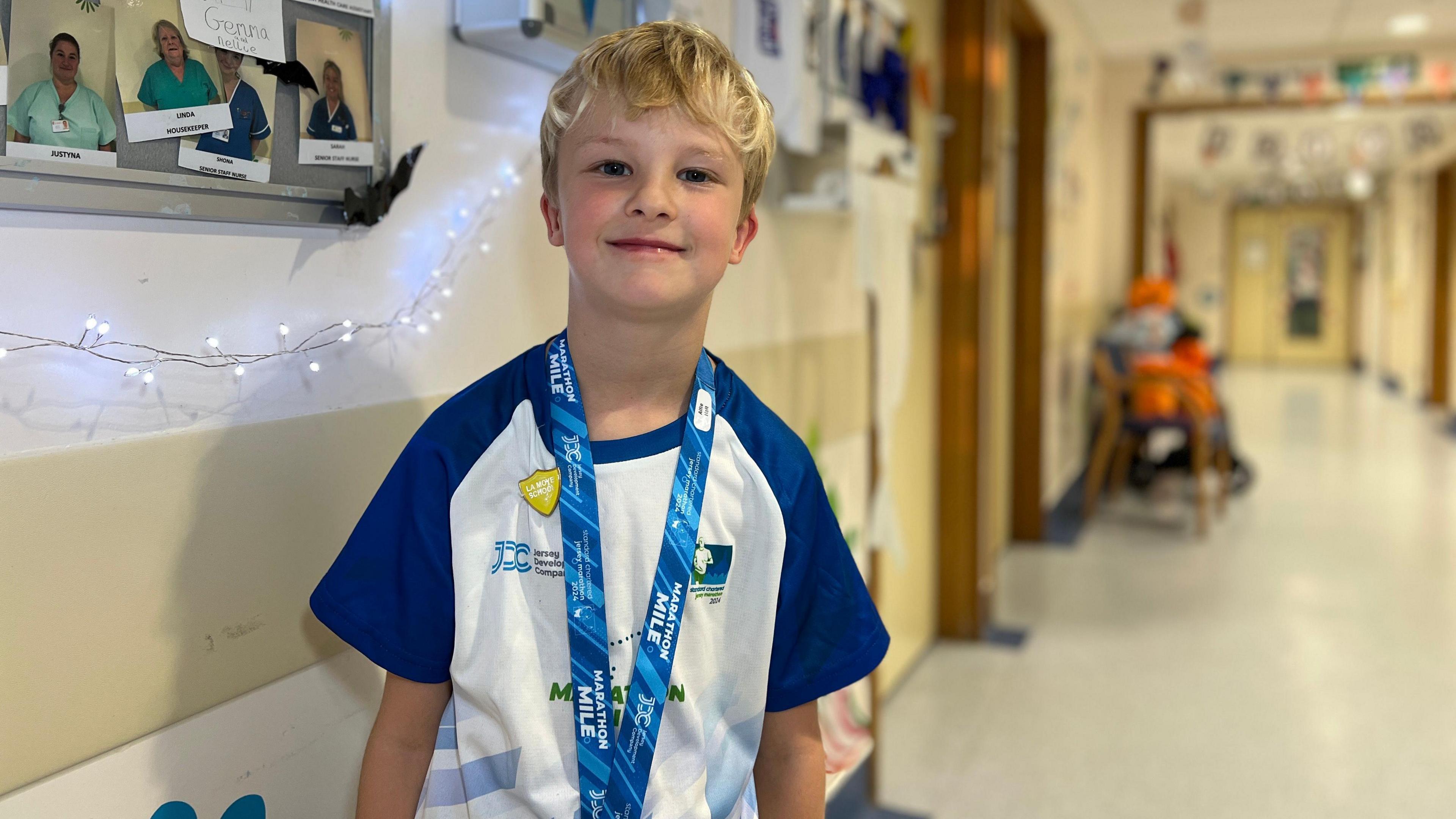 Alfie smiles at the camera as he stands in the hospital ward. He's wearing his running top from the Marathon Mile which has navy blue sleeves with a white middle. Alfie is wearing his medal around his neck too and a school badge as well. He has blonde hair and in the background is the ward corridoor.