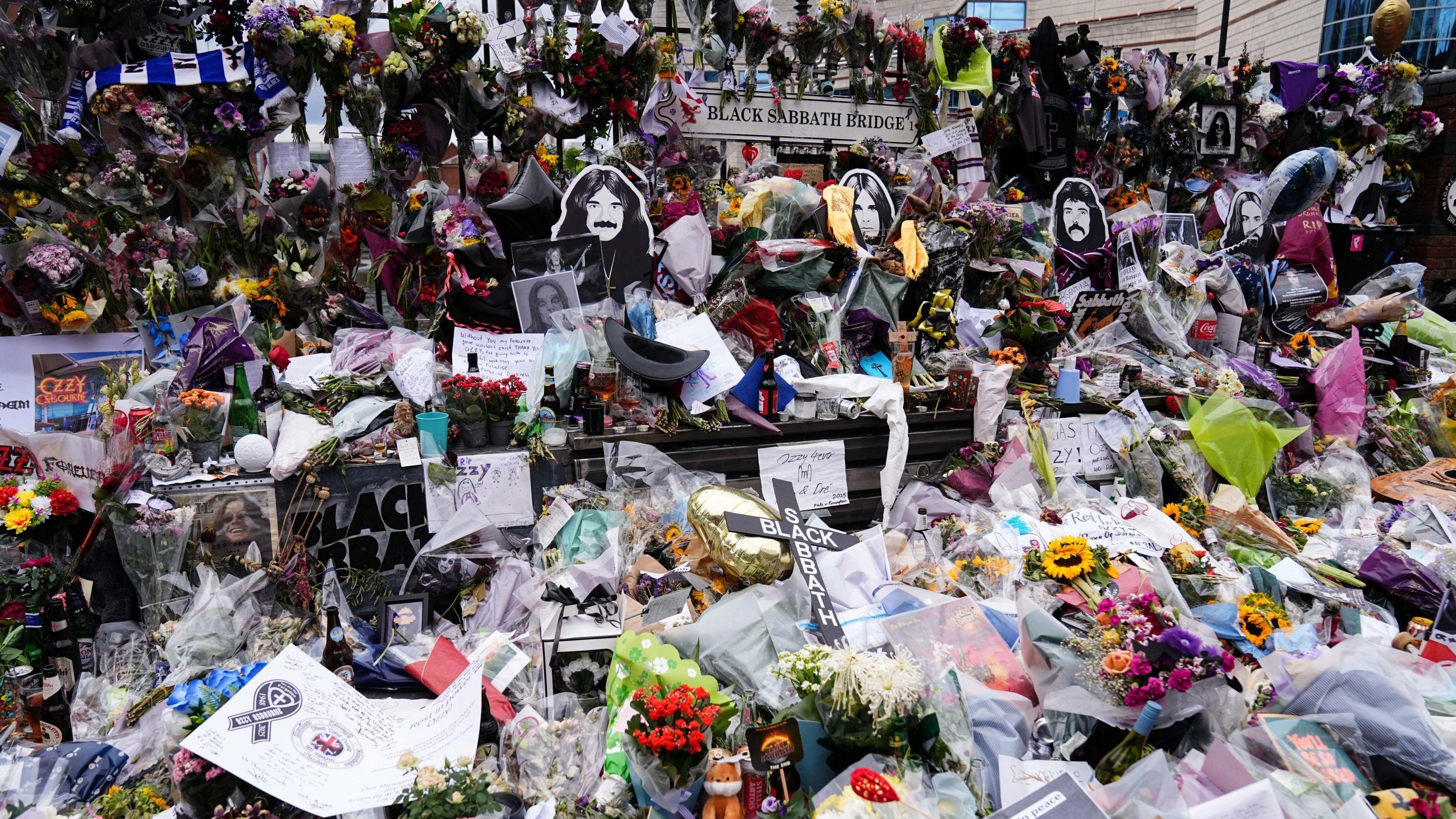 A huge pile of bunches of flowers, messages and other items left in tribute on a bench. Behind the bench are four cut-outs featuring each face of the Black Sabbath members. Behind them is a sign that says "Black Sabbath Bridge".