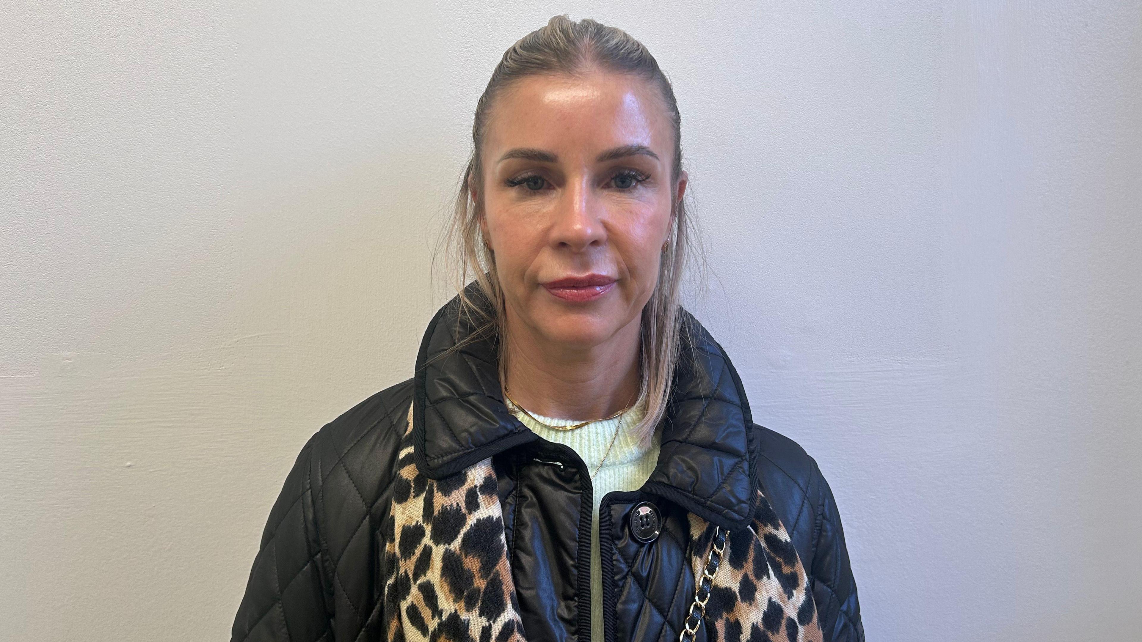 A women with light brown hair and red lipstick stands against a white wall. She looks into the camera wearing a black faux leather jacket and leopard print scarf, the chain from a bag is over her shoulder, across her body. 