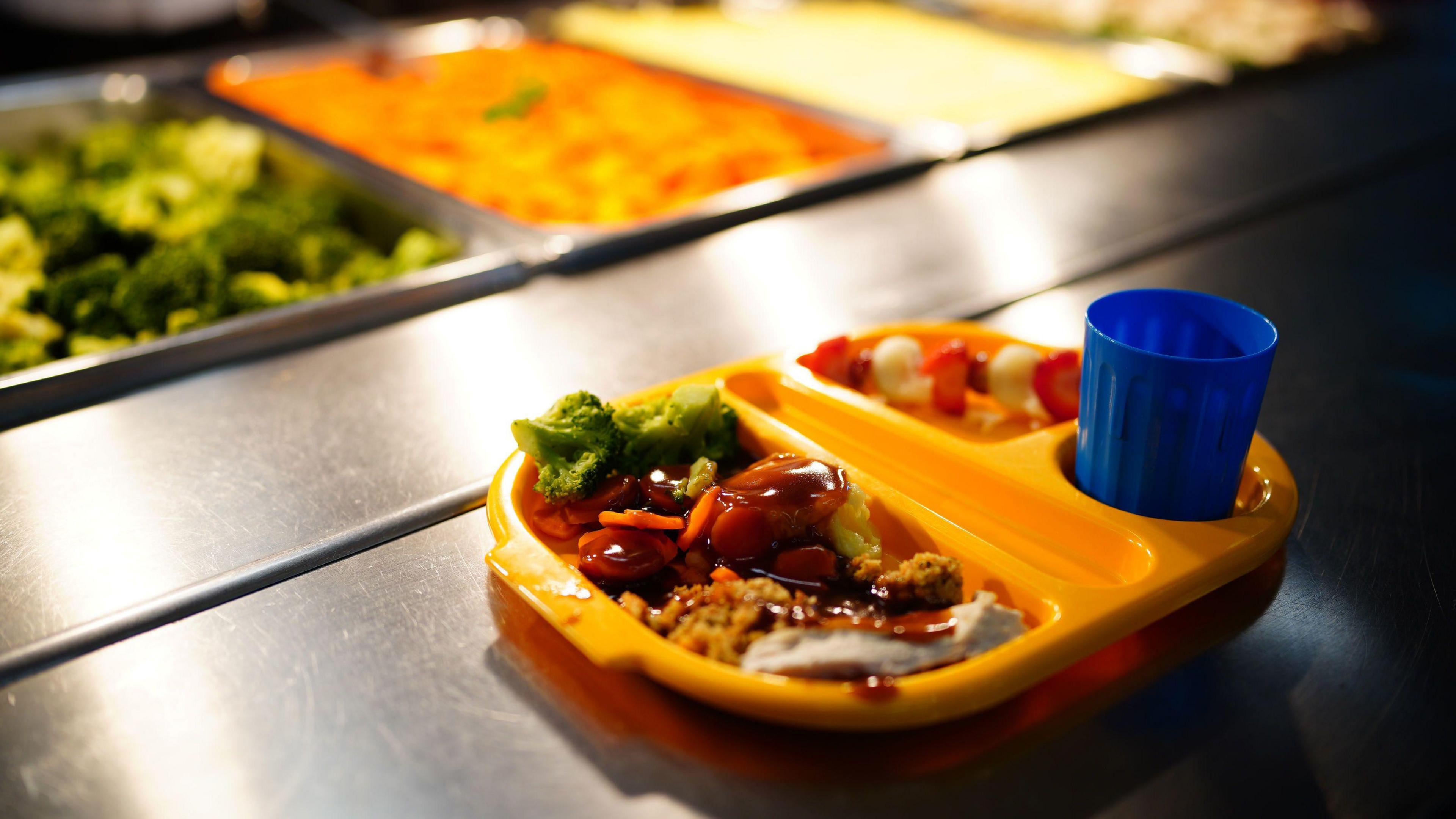 A school dinner containing meat and vegetables on a yellow tray with a blue cup. More food is also shown waiting to be served