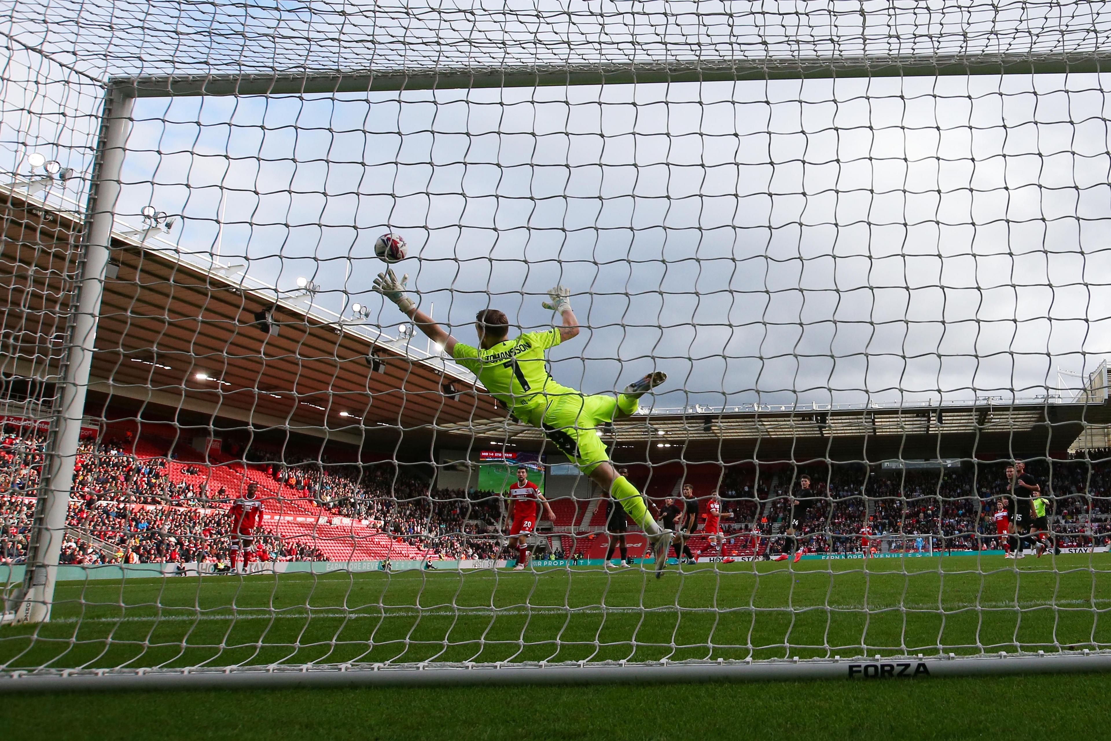 A goalkeeper in green dives to save a ball. The picture is taken from behind the net.