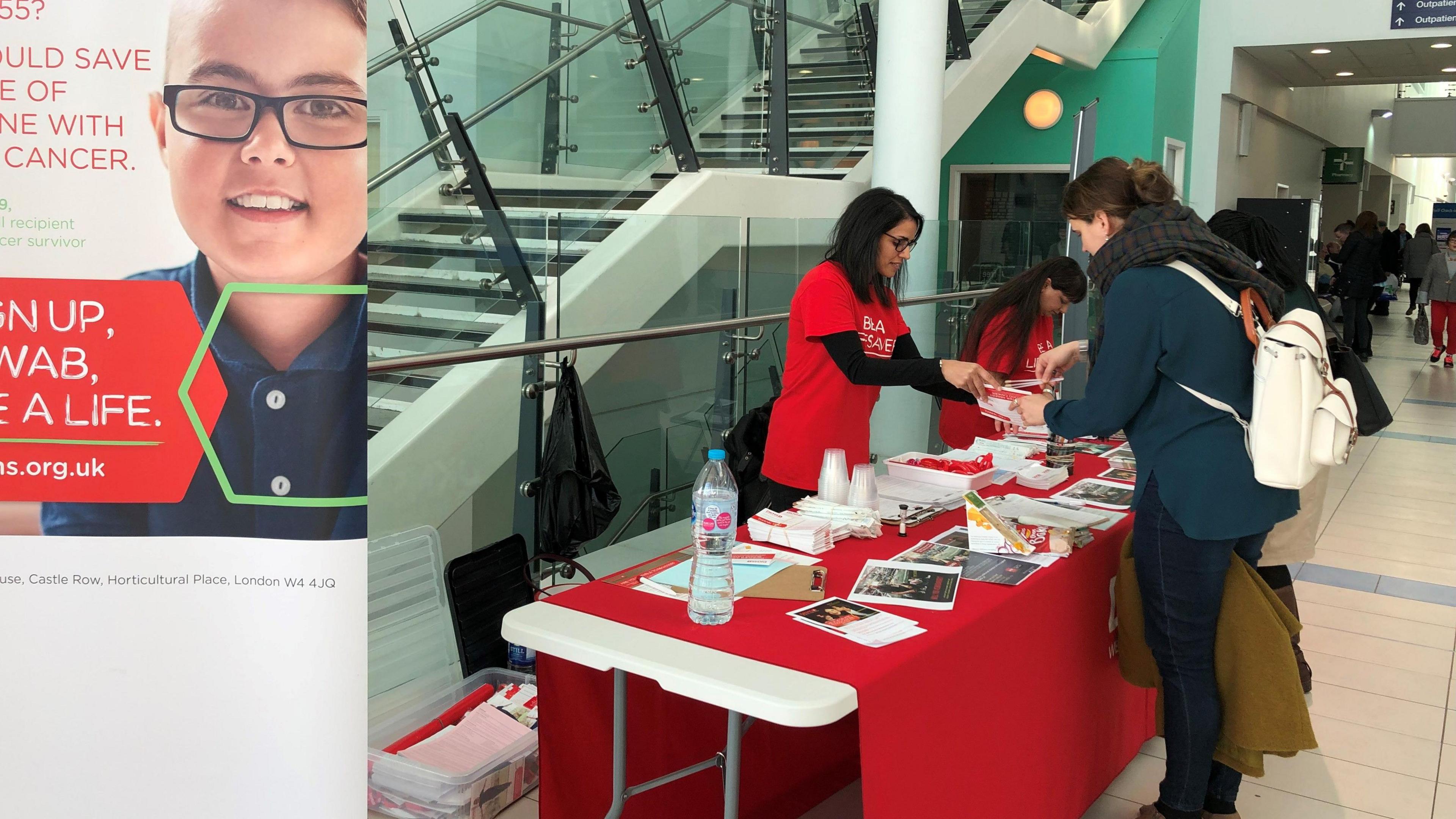 A woman with dark hair and glasses is behind a white table covered in a red tablecloth. She is wearing a red T-shirt. The table has lots of leaflets on it. She is handing a leaflet to a woman across the table.