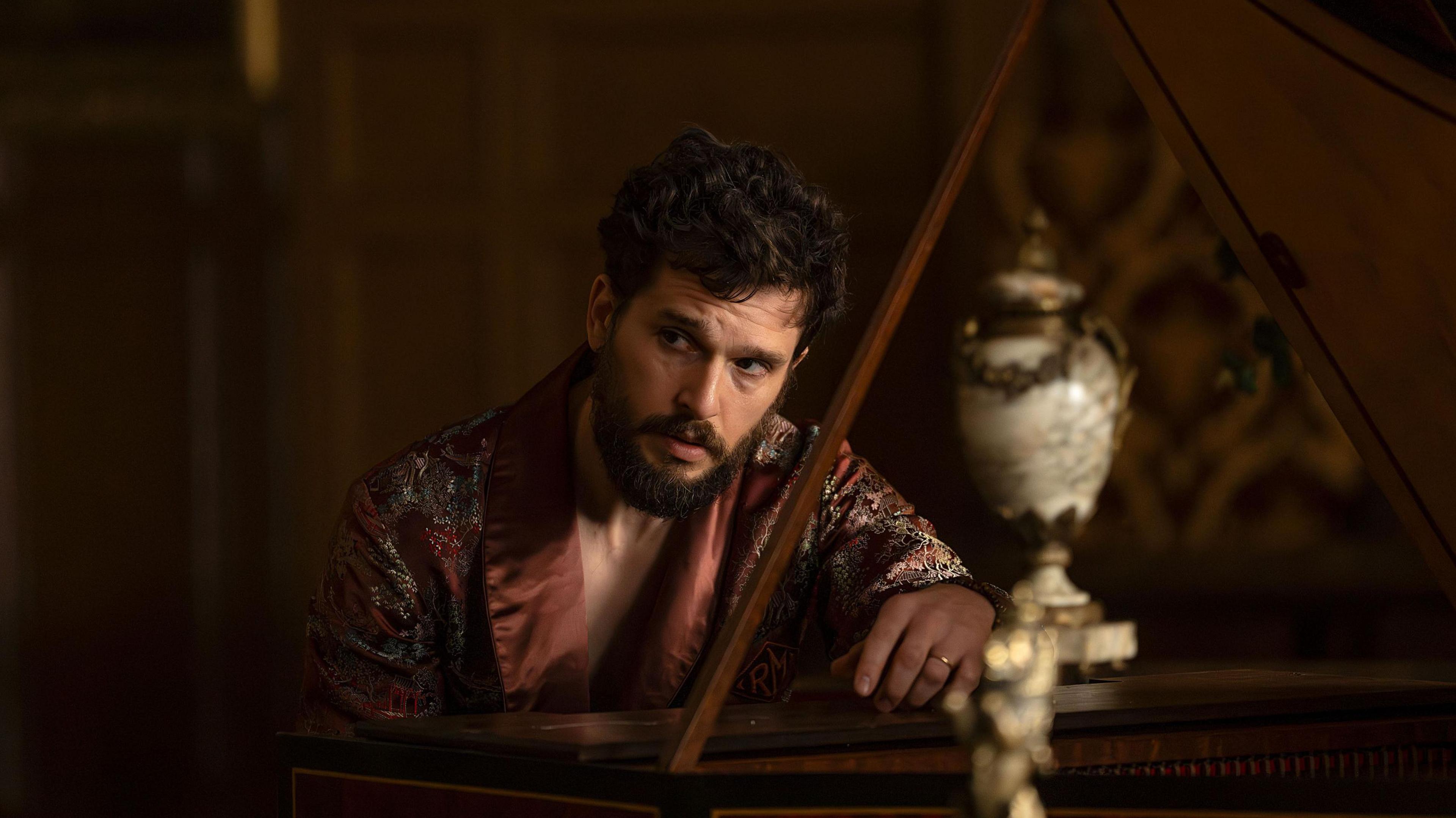 Kit Harington as Henry Muck, wearing a silk dressing gown, leaning over a grand piano