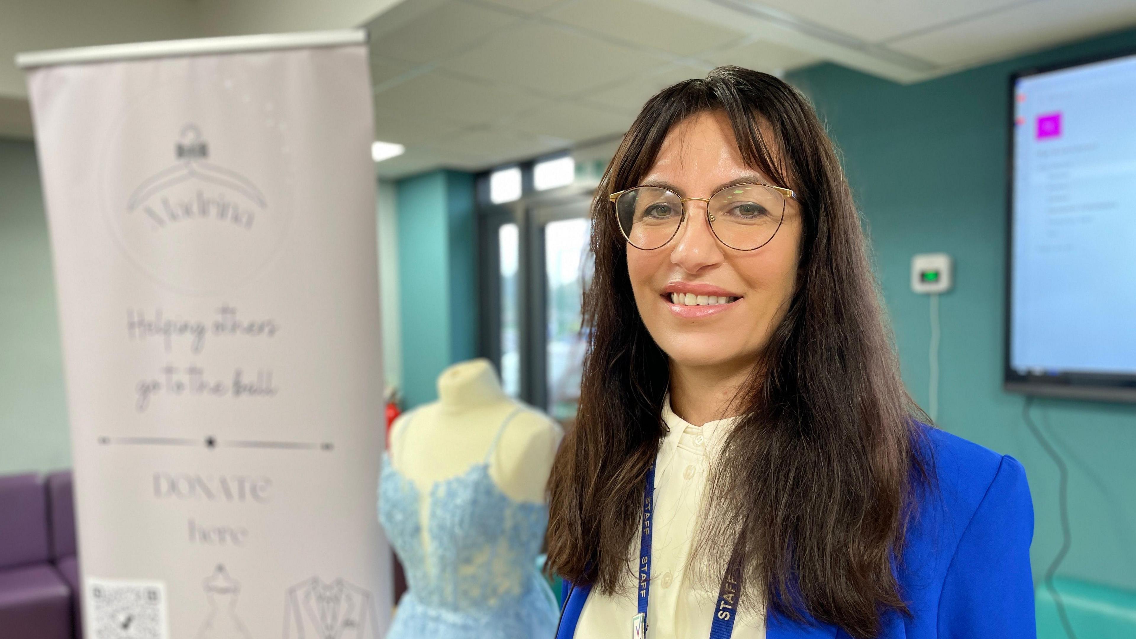 Stefanie Lakin stands in front of a mannequin which has a blue prom dress on it. To the side is is a pull-up stand with her charity, Madrina's, logo on it. She is wearing thin-rimmed glasses, a bright blue blazer, a cream shirt and a blue staff lanyard.