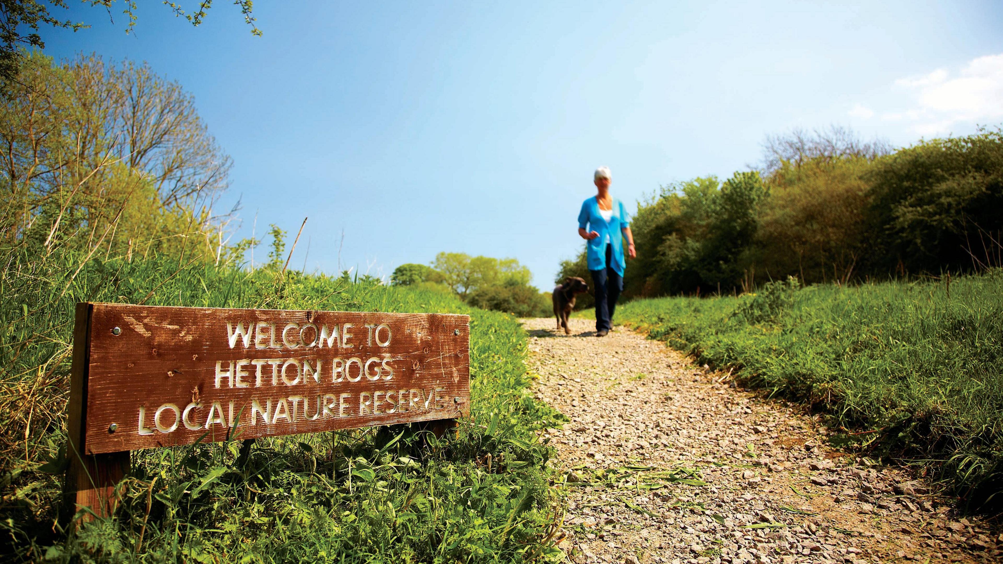 A small wooden sign reading "Welcome to Hetton Bogs Local Nature Reserve", sitting next to a path. A woman is walking along the path, towards the camera, with a dog.
