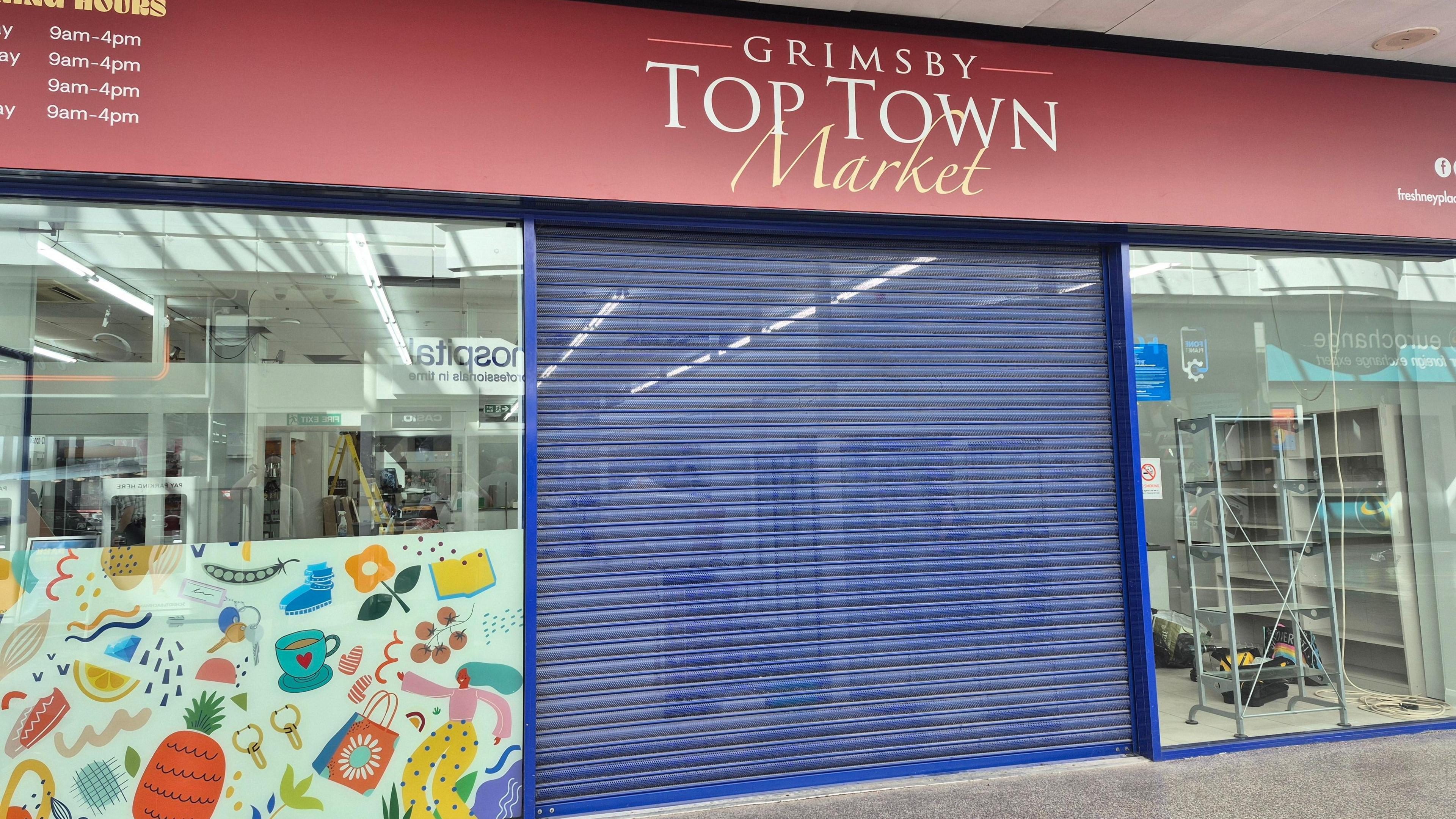 A shop unit with floor to ceiling glass windows either side a blue shuttered entrance. Above is a burgundy sign with 'Grimsby Town Town Market' in white lettering in the middle, on the left are opening times also in white type face.