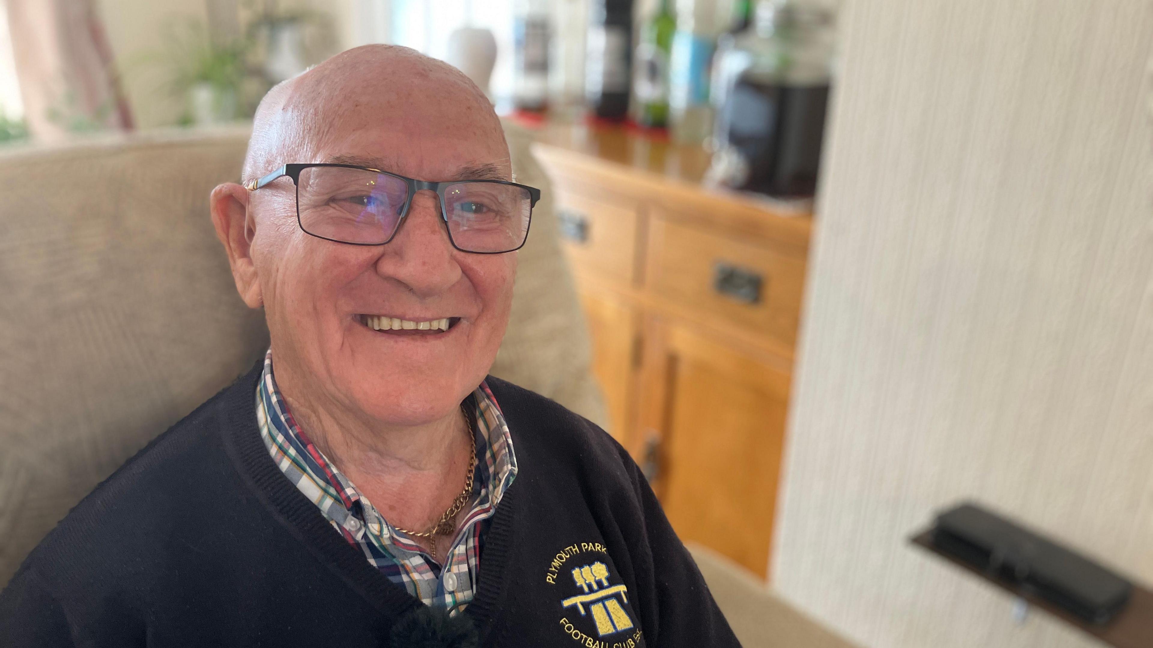 A close up of David Liddicoat at in a cream arm chair. He is wearing a Plymouth Park Football club navy blue jumper, he is smiling and is wearing black glasses. The background behind him is blurred and appears to have a cabinet with plants on it.