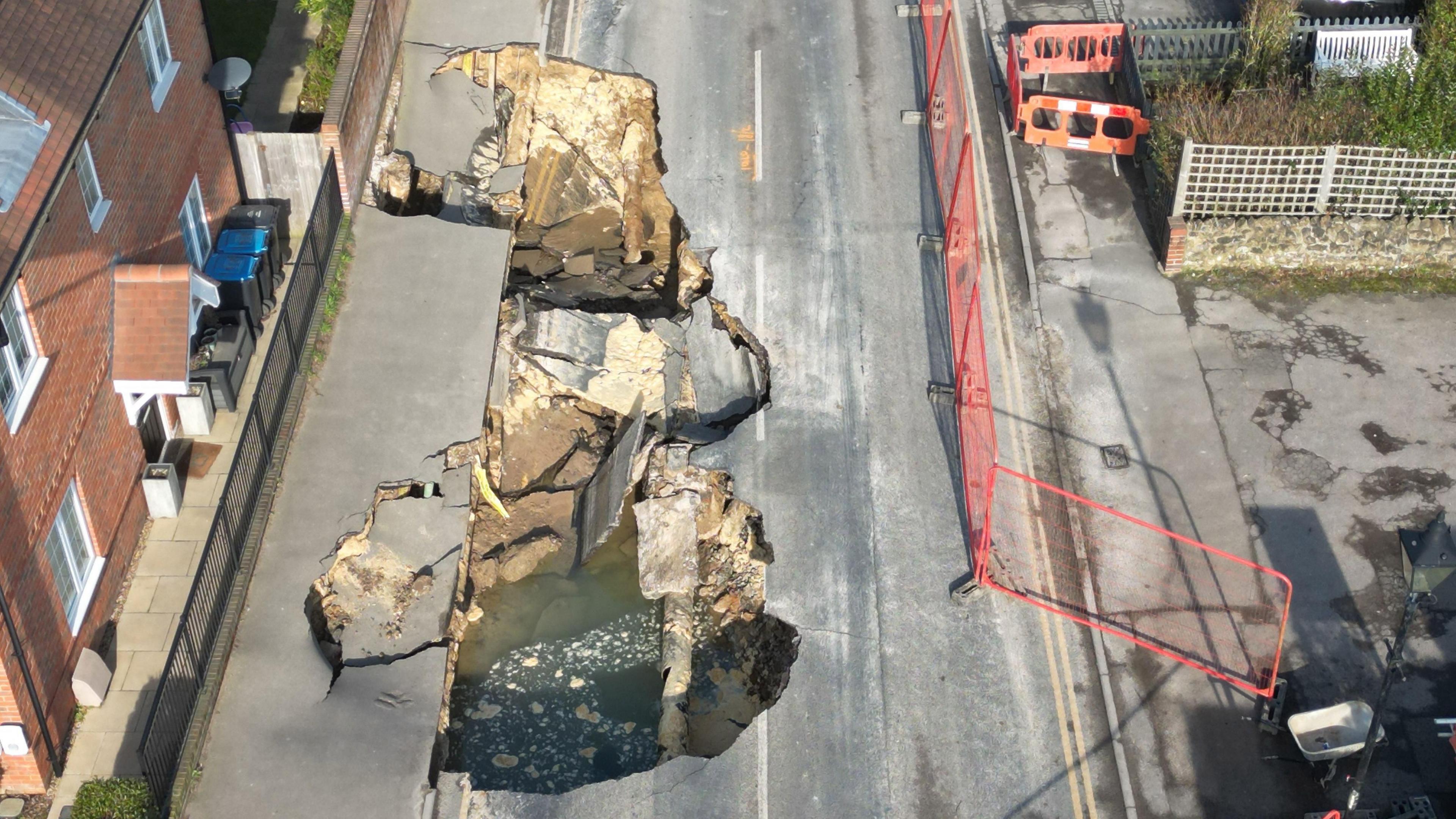 An enourmous hole in a road full of mud, water and road debris beside some red brick houses seen from the air. 
