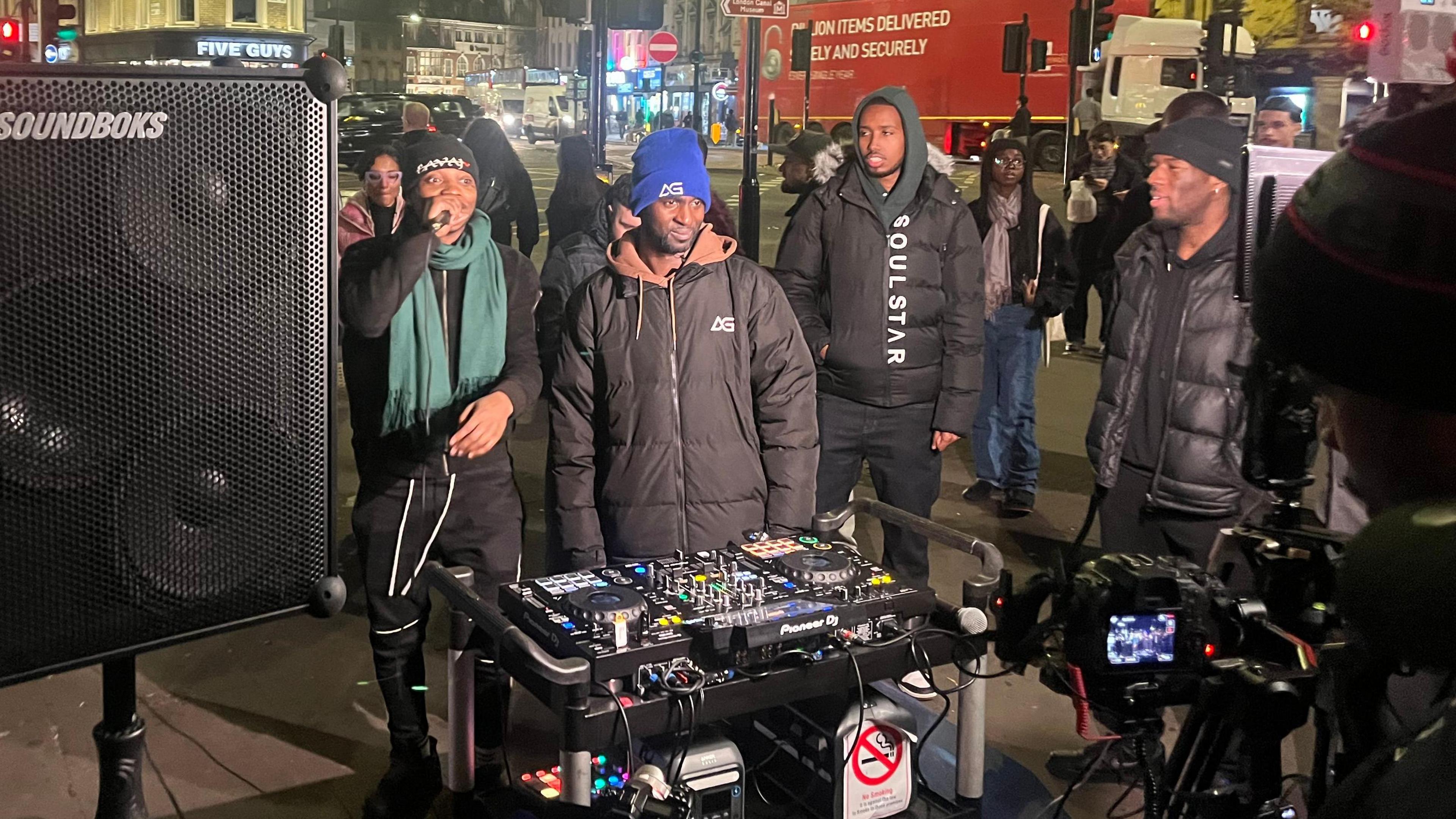 DJ decks on a trolley with a big speaker on the left. There's a camera in front of the DJ as he goes live on Tik Tok. Behind the decks, there's a man with a blue had, brown hoodie and black puffer jacket with the logo AG on it. He's flanked by a man in a gree scarf rapping  as well as onlookers. 