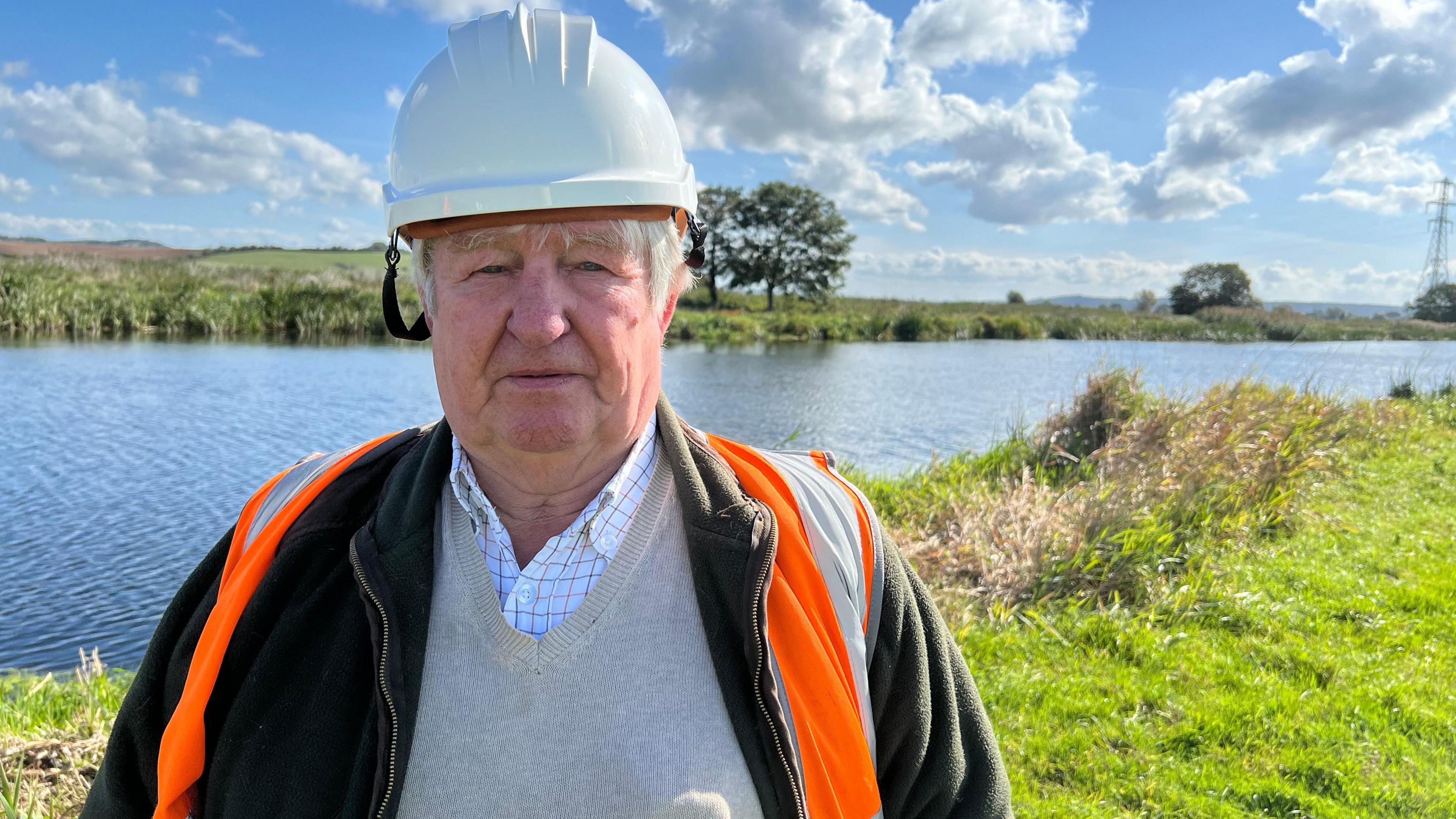 Tony Bradford is standing by a river wearing a white hardhat and an orange high-vis vest. The river is blue and there is green grass next to it.