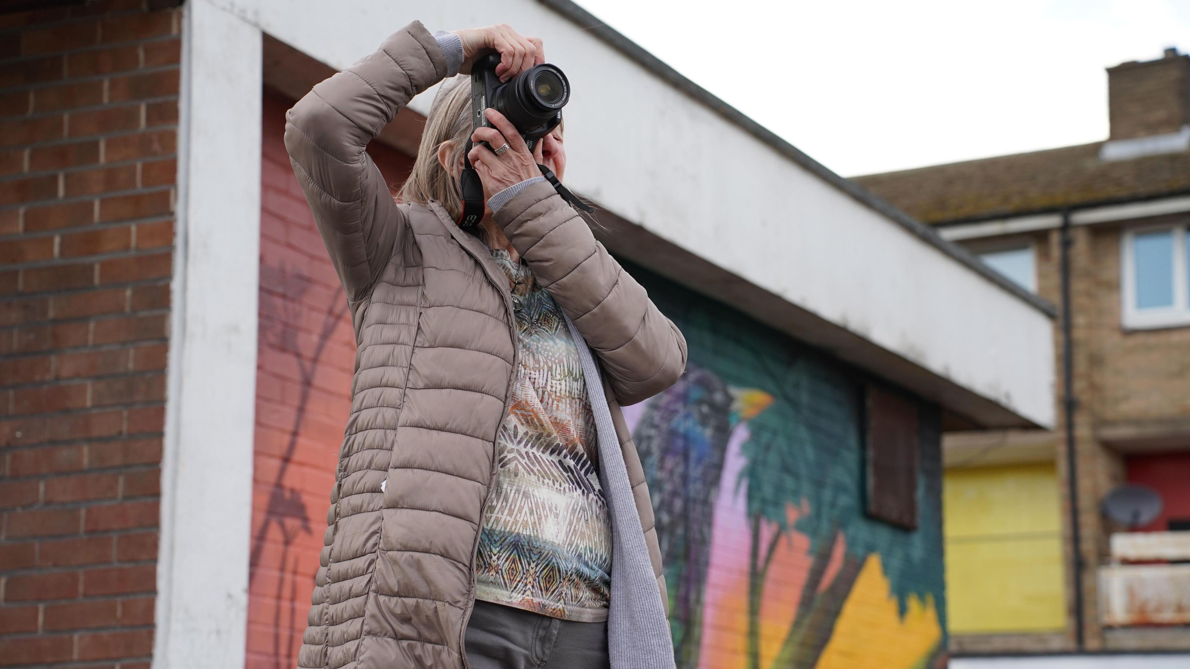 A woman with short blonde hair in a beige puffer jacket takes a photo through her camera. Behind her a brightly coloured jungle scene is painted on brickwork.