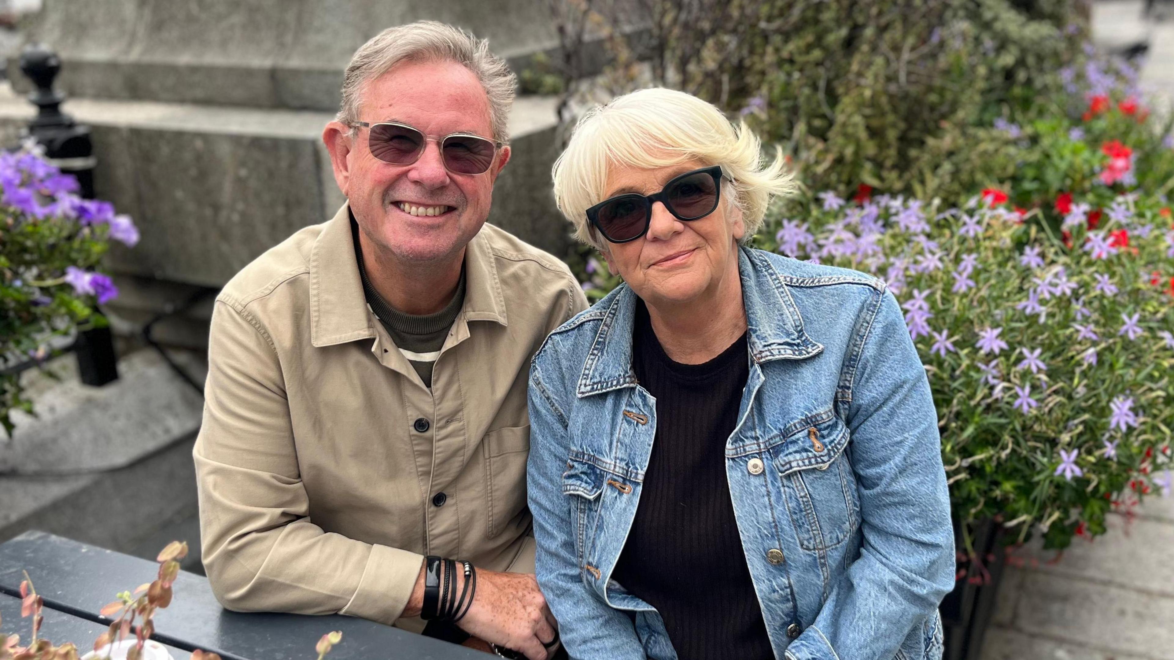 Chris and Martine Dean are sitting on a bench in St Peter Port in front of some flowers. Chris is on the left wearing sunglasses and a thick, collared brown shirt. Martine is on the right and wearing a denim jacket with a black top and sunglasses. 