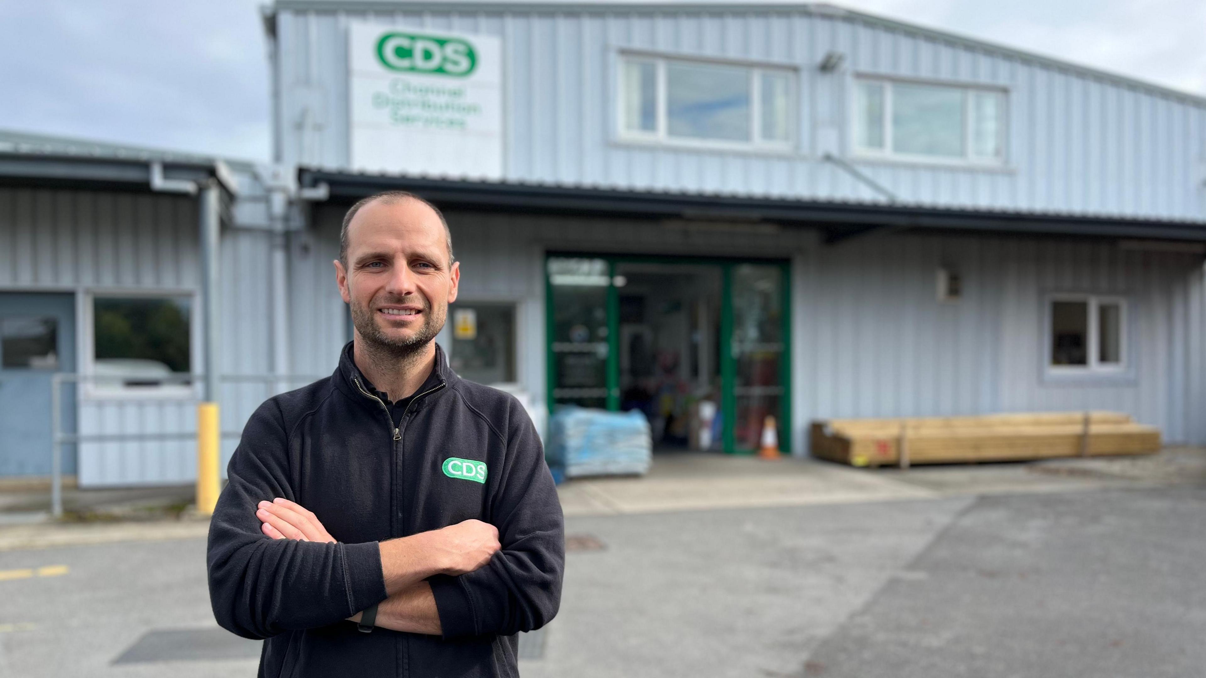 A man in a black jumper with crossed arms outside a builders merchants.