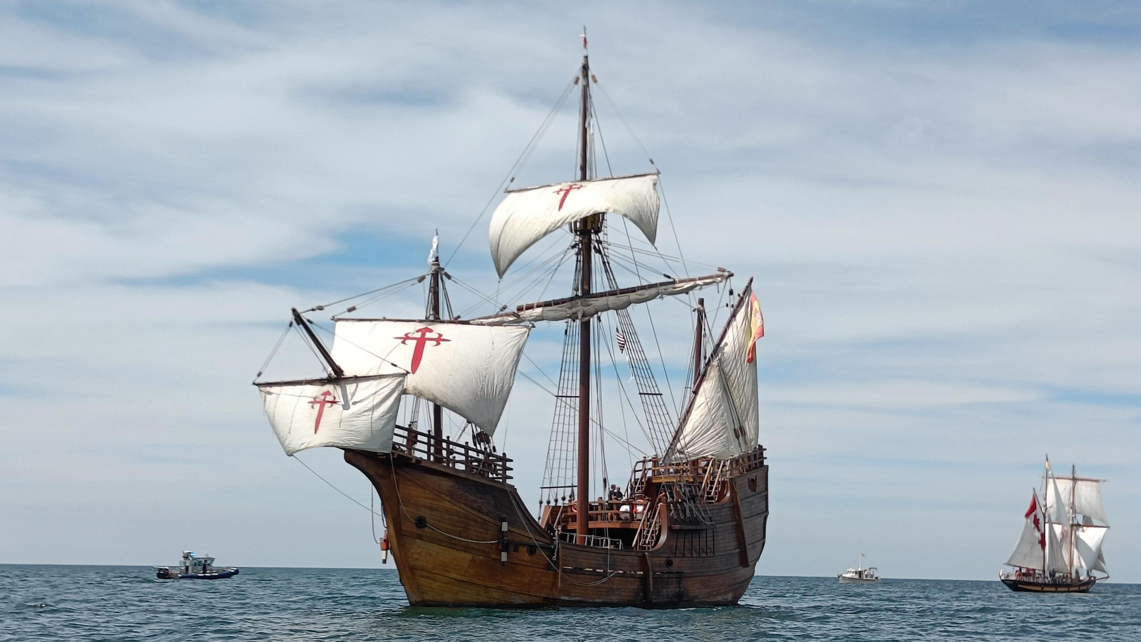 A wooden ship in the ocean. It has large white masts with a red gross on them. There is a Spanish flag flying from the back.