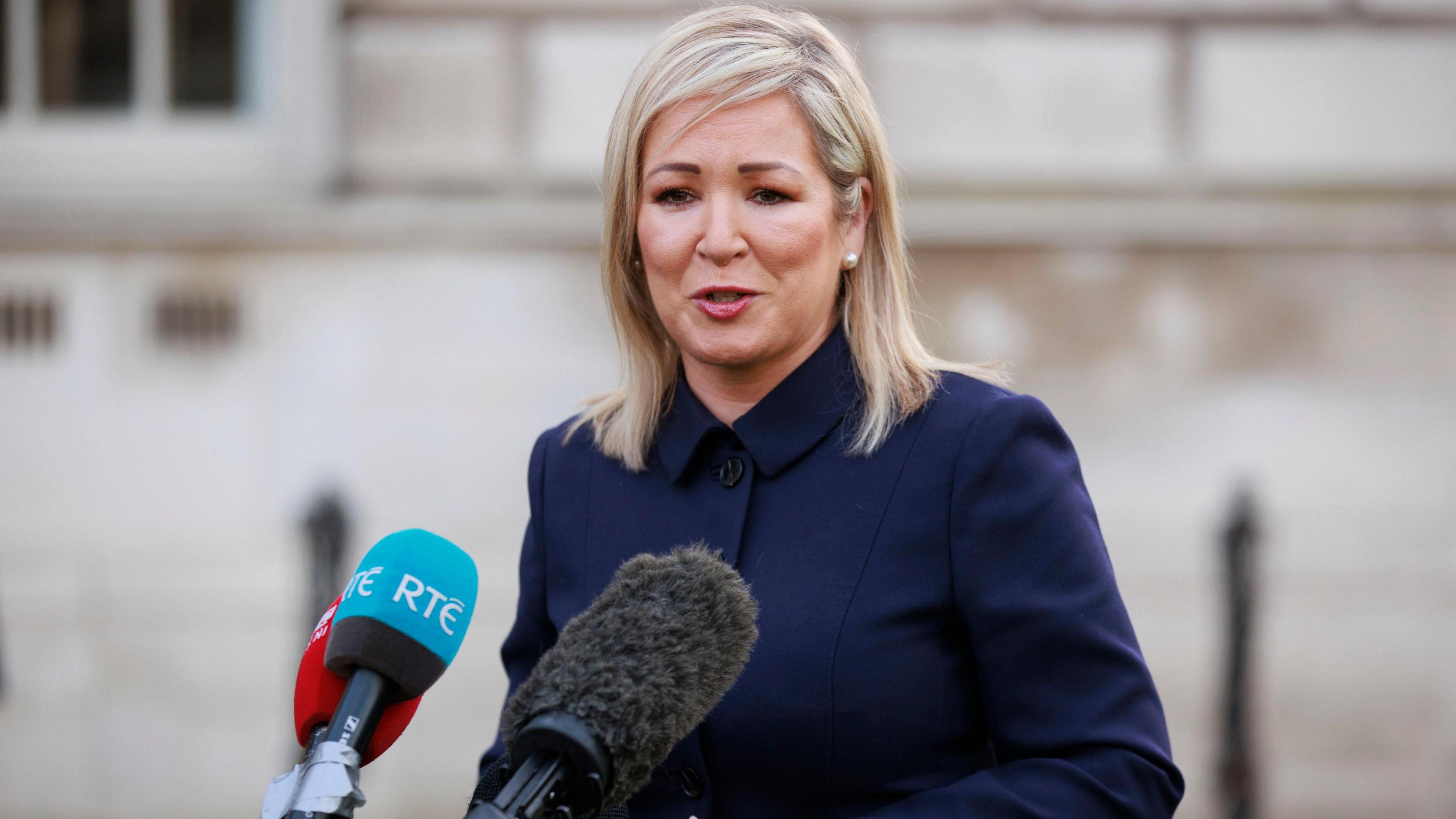 Michelle O'Neill stands in front of three microphones before a white brick building. She has shoulder-length blonde hair and is wearing a navy coat.