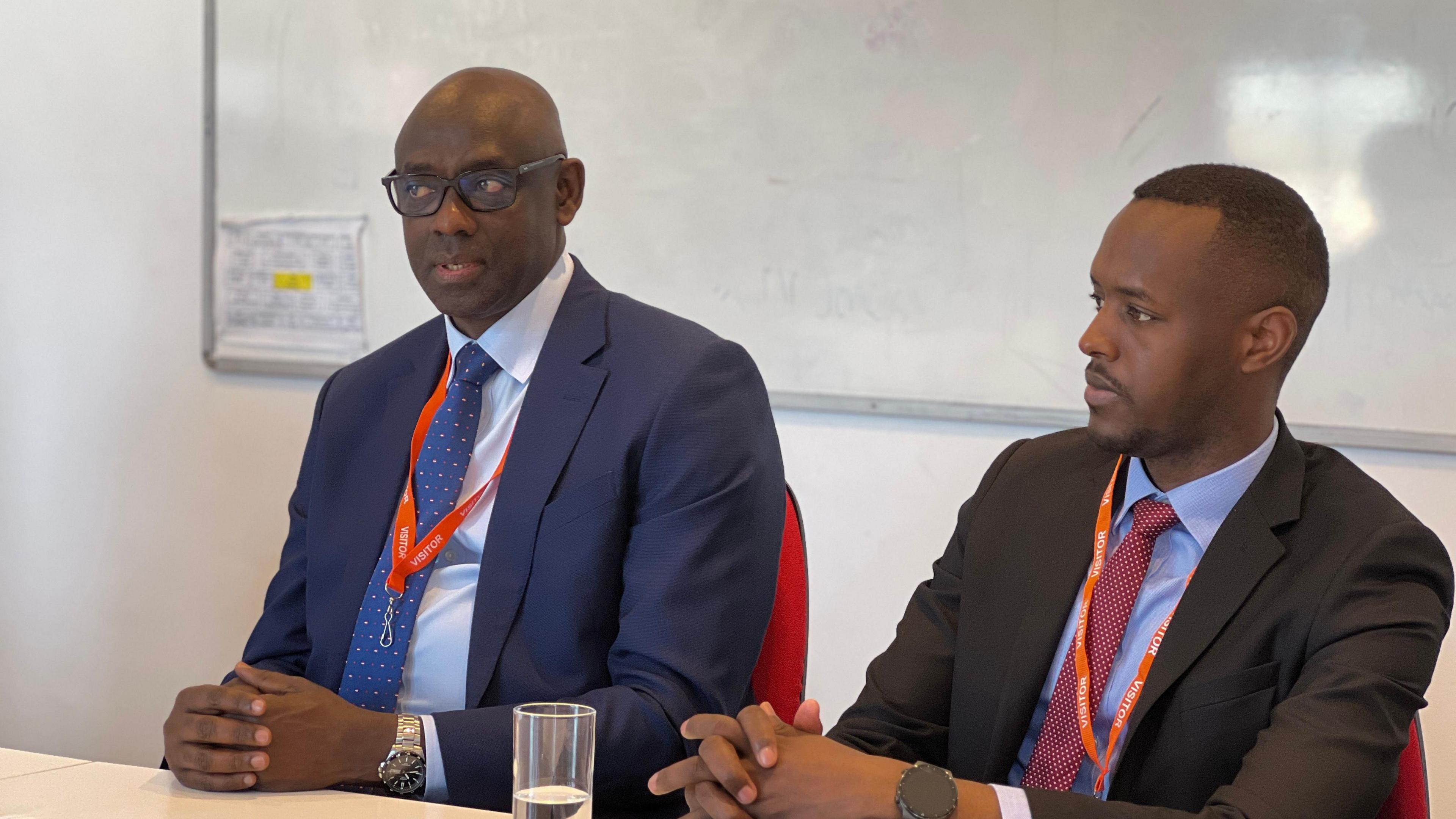 Johnston Busingye  sits in front of a white board sat in front of a desk and another man is sat to his right