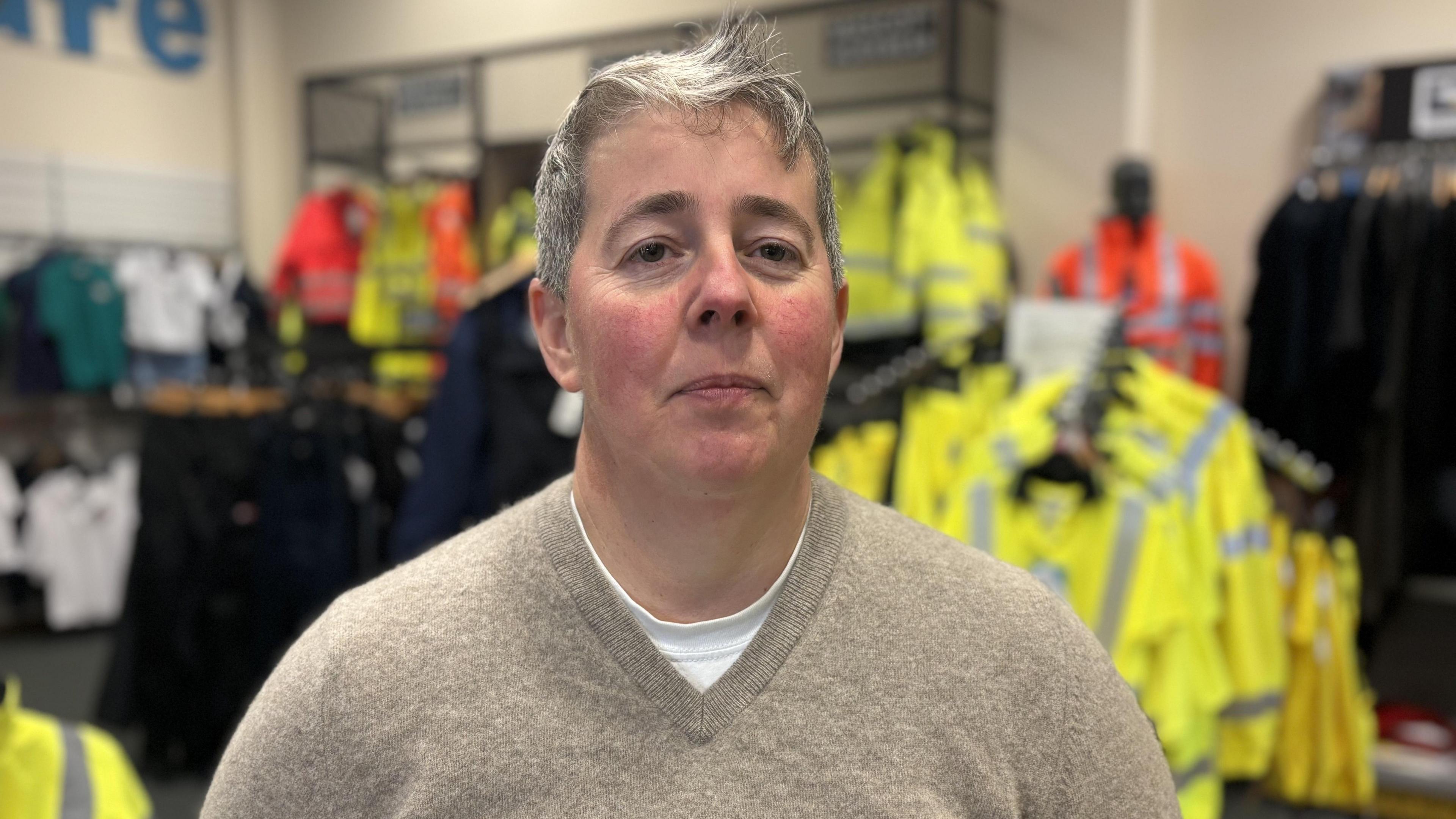 Mae Rhian Parry yn edrych yn syth ymlaen, mae'n gwisgo siwmper olau a chrys T gwyn oddi tano. Tu ôl i'w phen a'i hysgwyddau mae dillad gwaith lliwgar yn hongian.