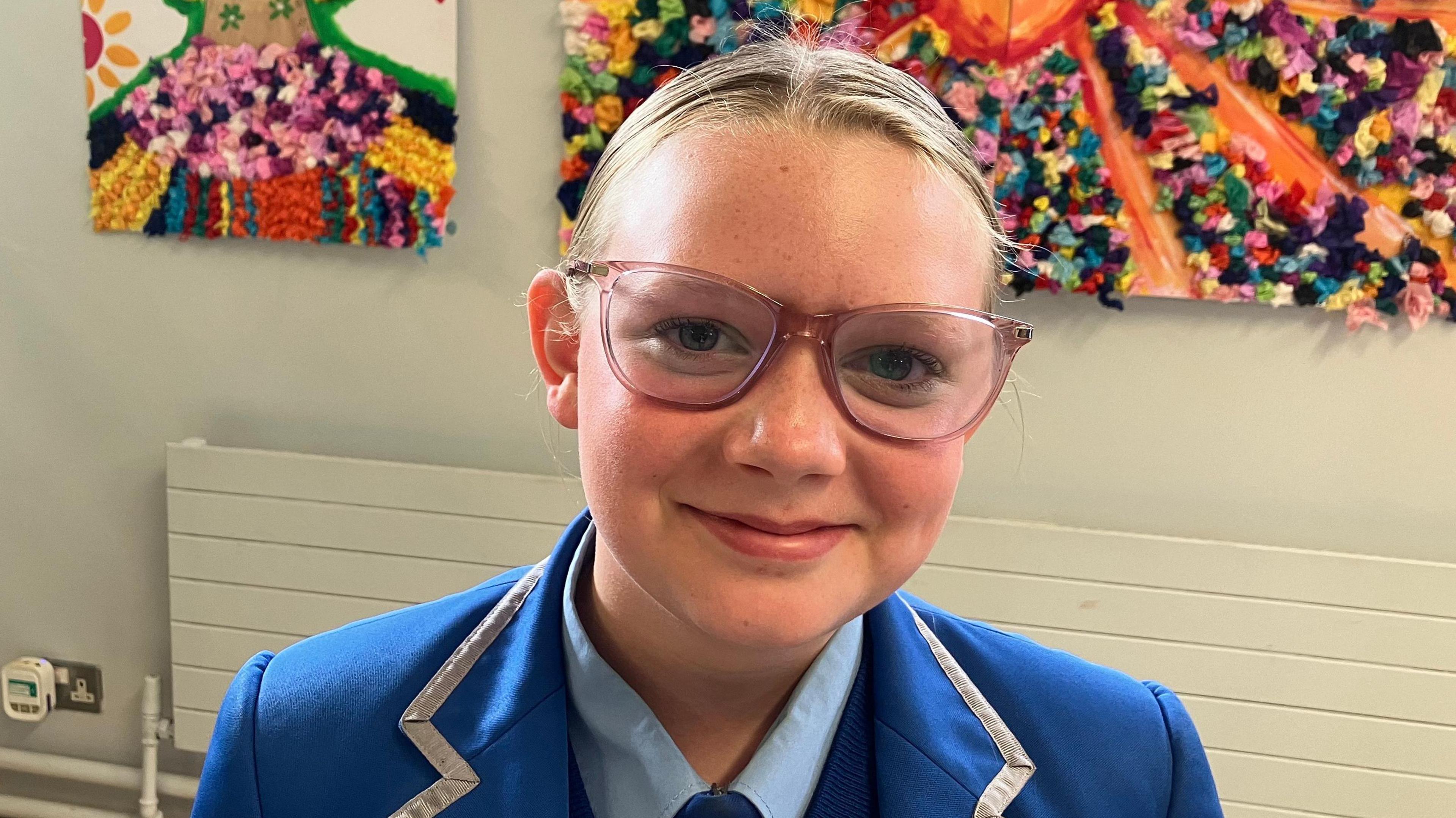 A schoolgirl is smiling into the camera. Her uniform is blue. She's wearing glasses. Art is hangin on the walls behind her.