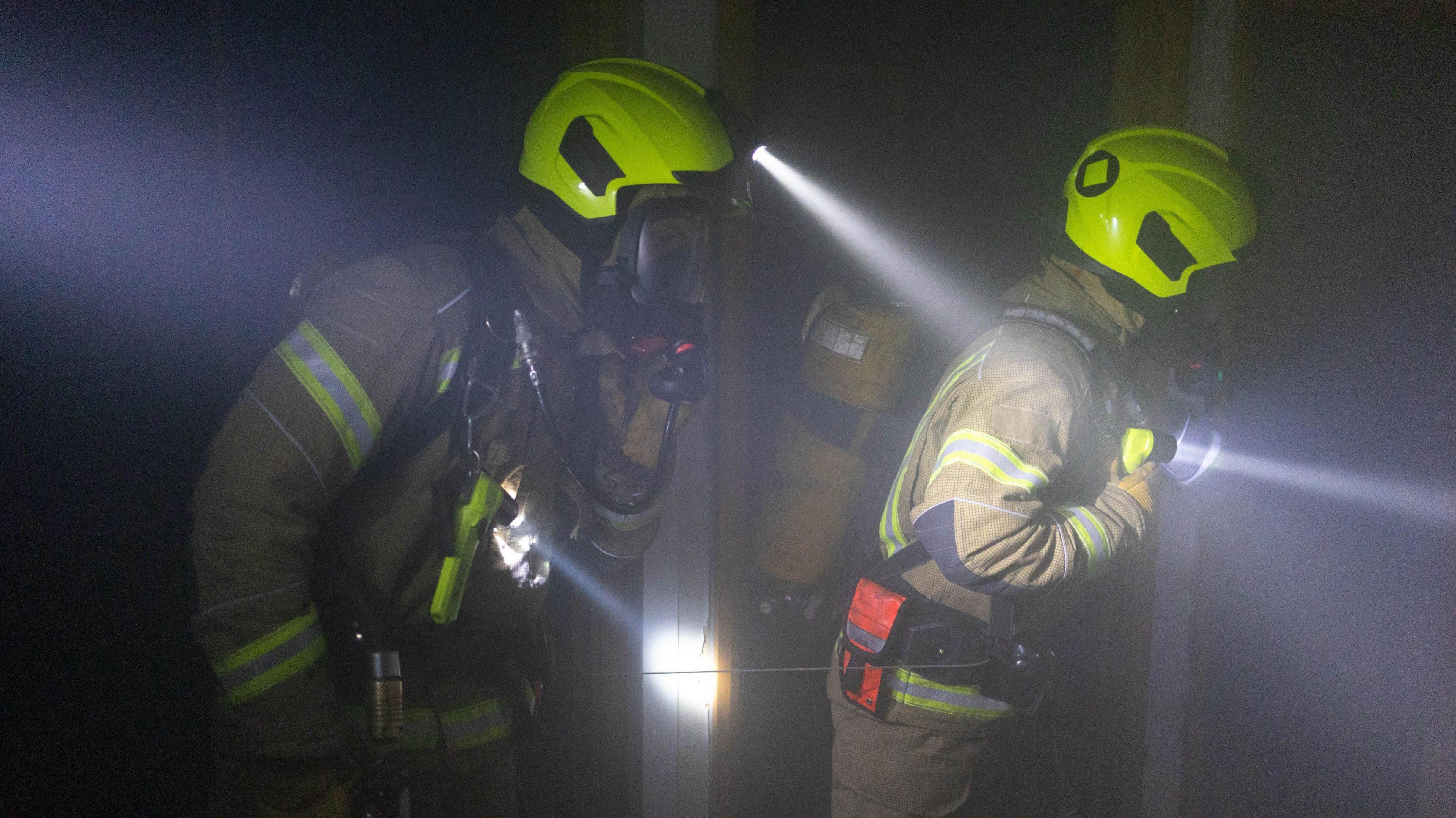 Two firefighters with torches inside a smoke-filled building. 