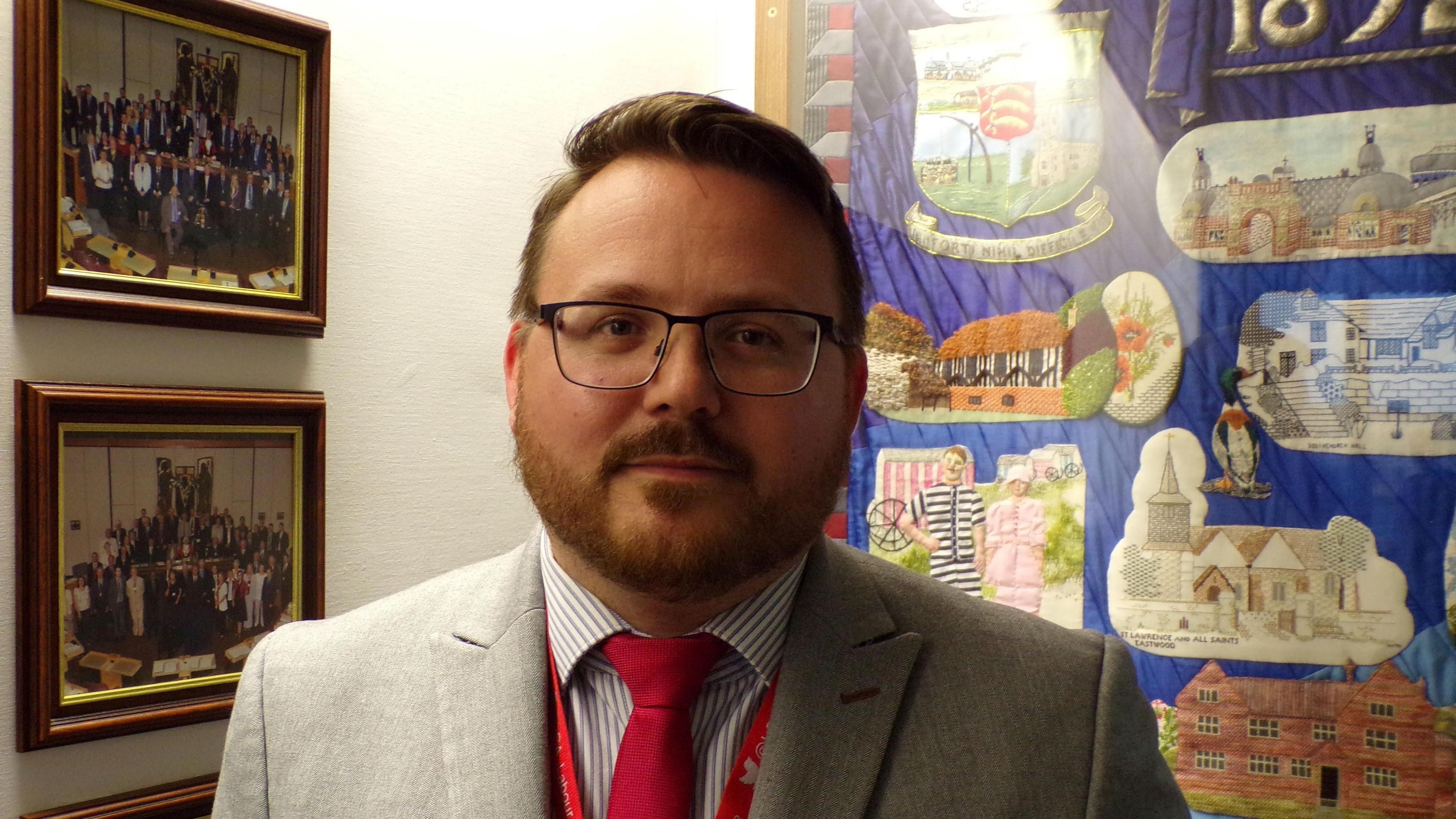 David Cowan, wearing dark framed glasses and with light brown short hair and short beard, outside Southend's council chamber, standing in front of a blue fabric wall hanging, covered in appliqued buildings