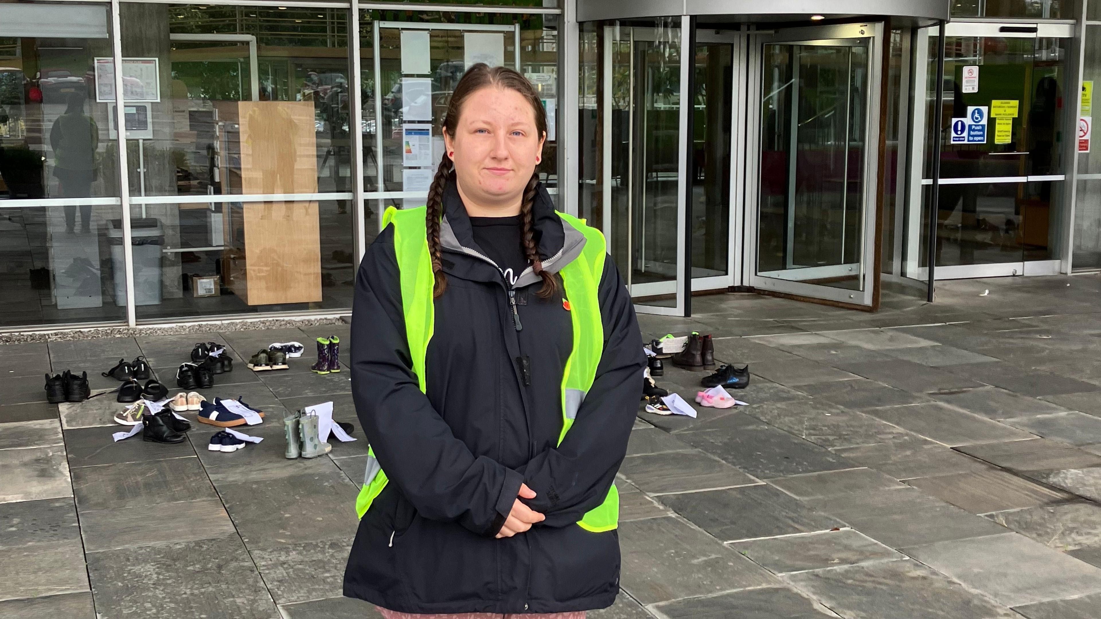 Louise Gilburt is a woman with mid-length brown hair in braids. She is wearing a black coat and green high-viz jacket, again standing in front of County Hall. On the grey concrete behind her are children shoes. 