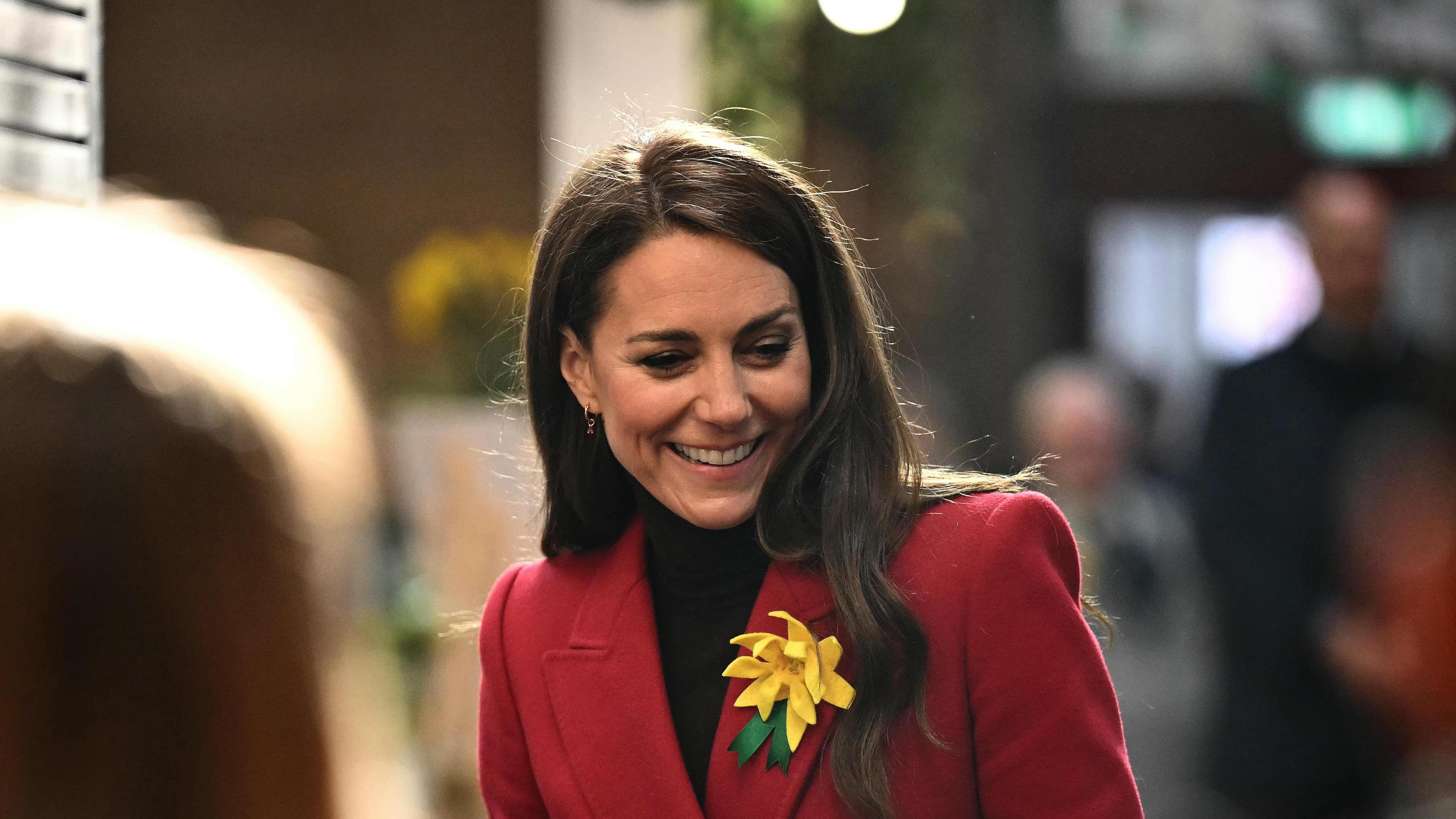 The Princess of Wales is smiling as she looks down, she is wearing a red coat and is holding a pink and white polka dot tube. Catherine has a large yellow daffodil broach attached to her red coat. She is in focus and the foreground is people waiting to meet her, their heads are out of focus.
