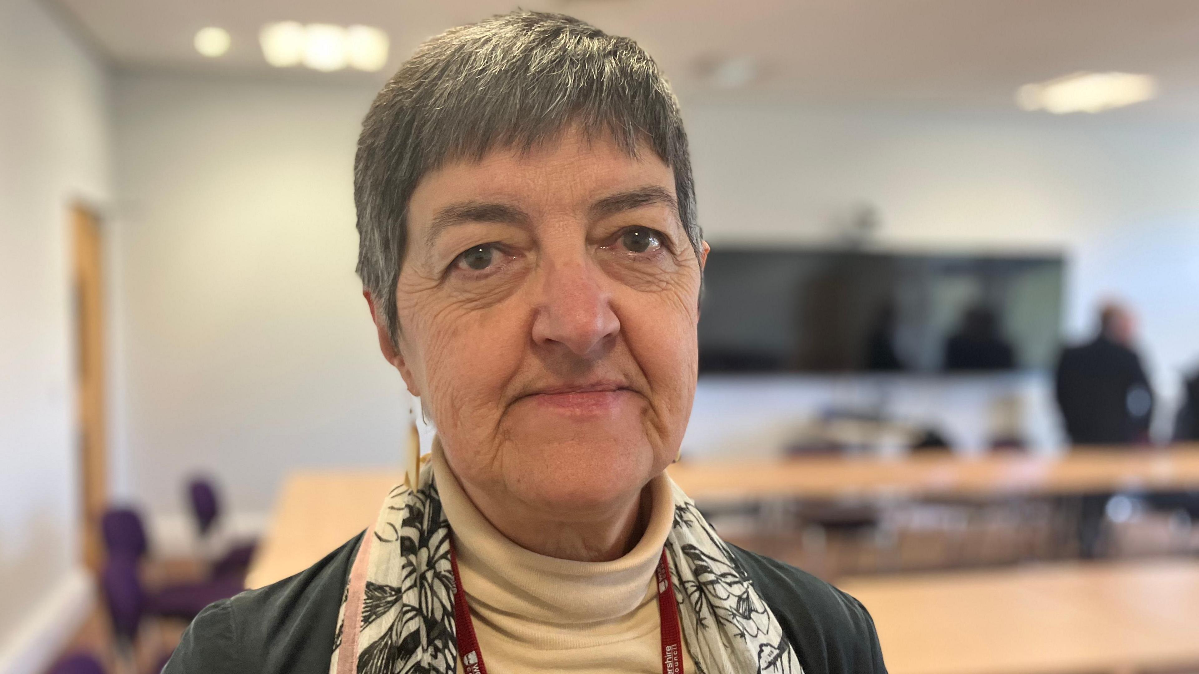 Council leader Lynn Denham in a close-up photograph. She is wearing a beige polo neck and scarf and jacket. She has short hair and is wearing earrings. She is in a council room with people behind her at a table.