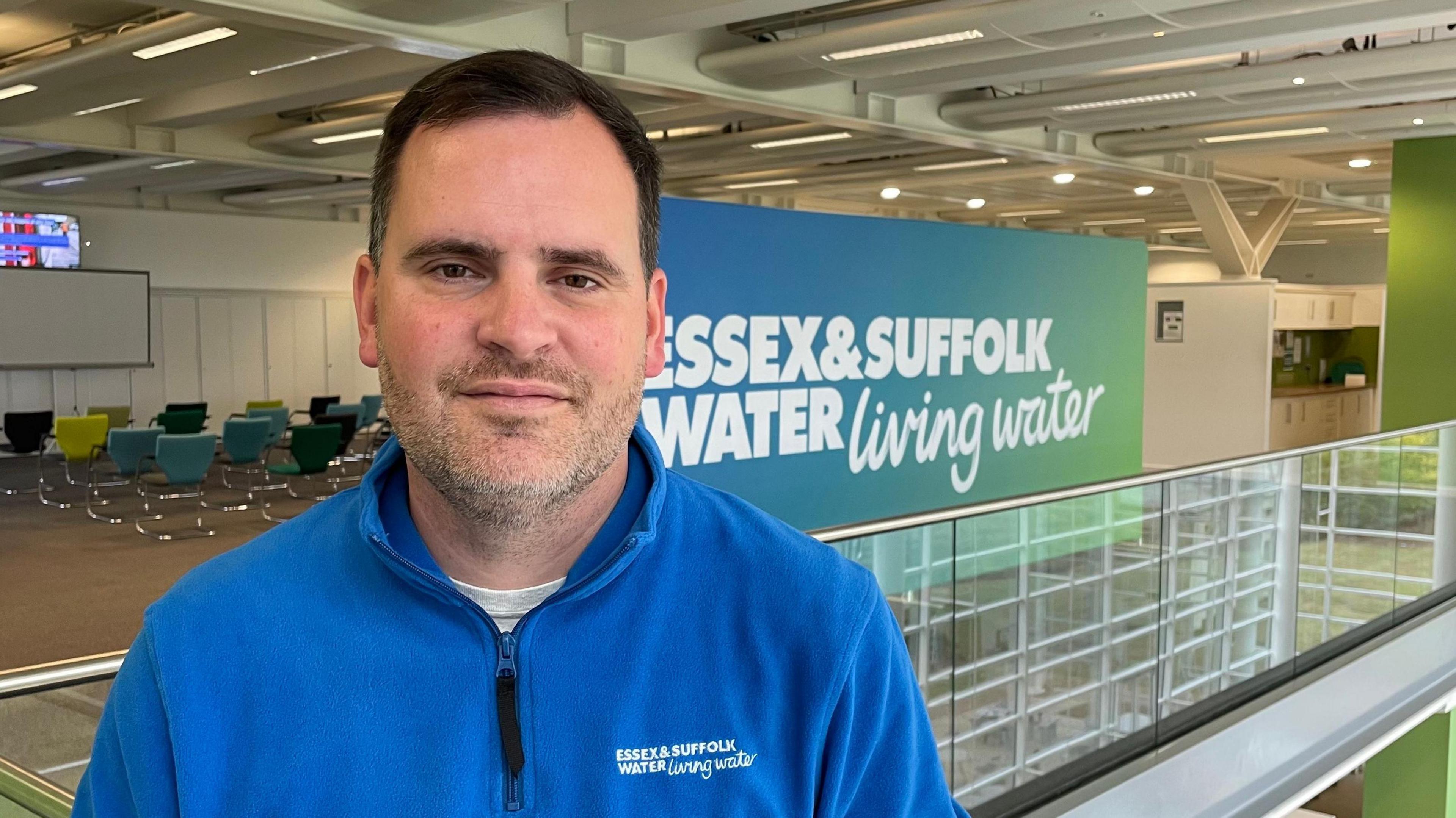 Kieran Ingram is looking directly at the camera, wearing a blue quarter-zip fleece with the Essex & Suffolk Water logo visible. He is standing in front of a large sign that also bears the company's name. He has dark hair and eyes and is unshaven. He is standing in an open-plan, indoor space. There are several rows of empty chairs visible over his shoulder.