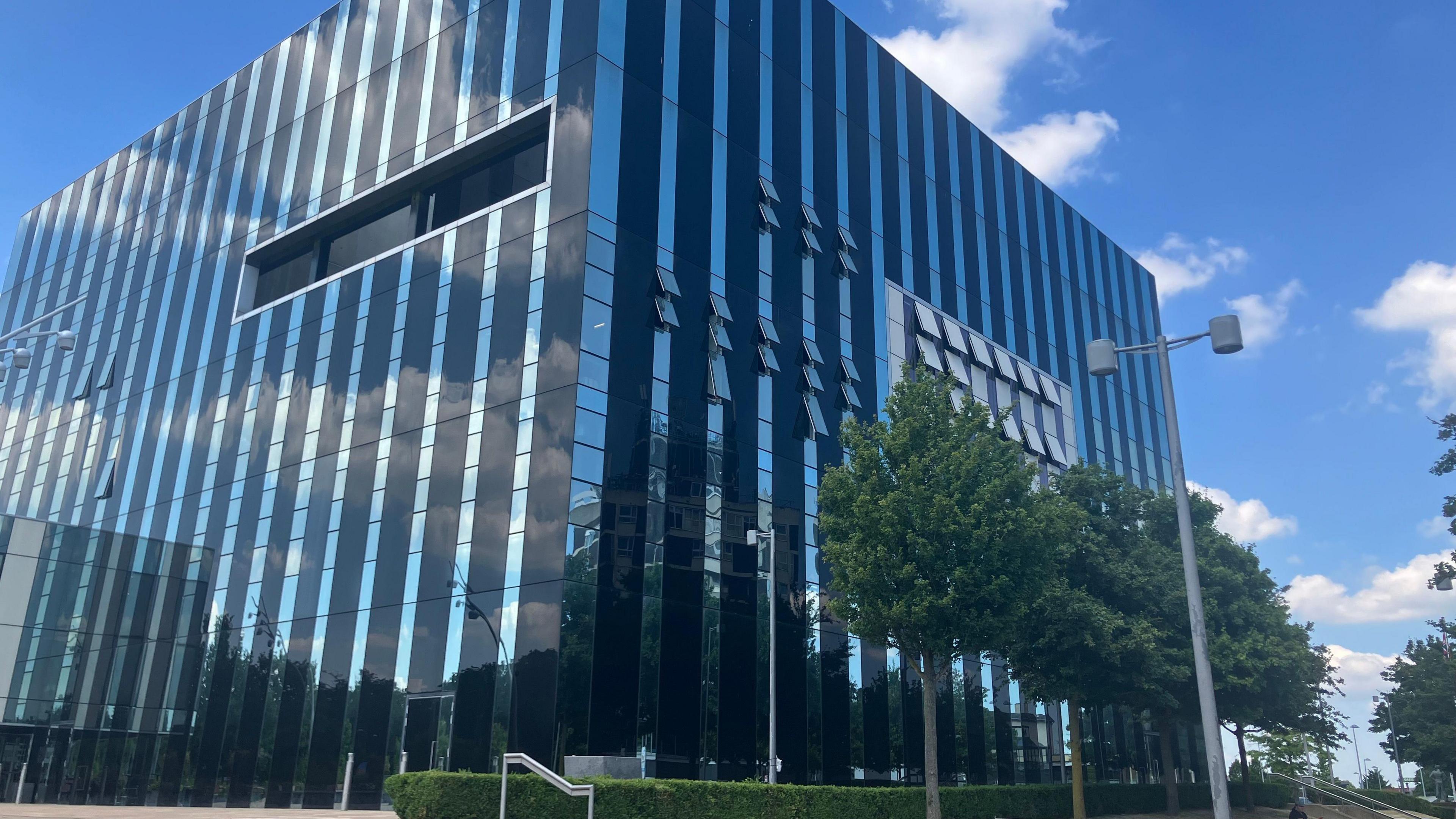 The glass and steel exterior of the Corby Cube building, headquarters of North Northamptonshire Council, on a blue sky day.