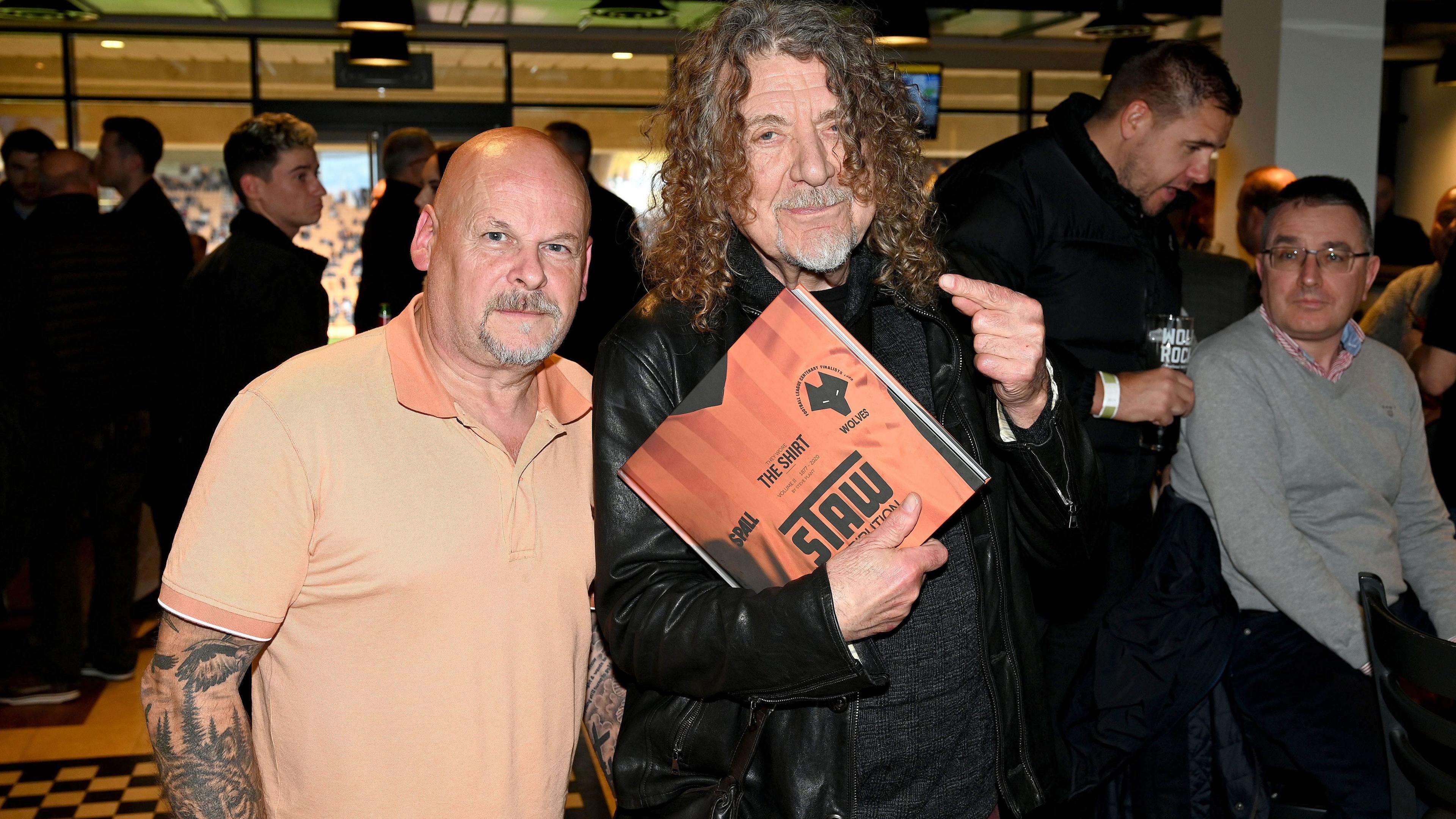 Two men, one bald, one with long curly hair and holding a copy of a book about football shirts 