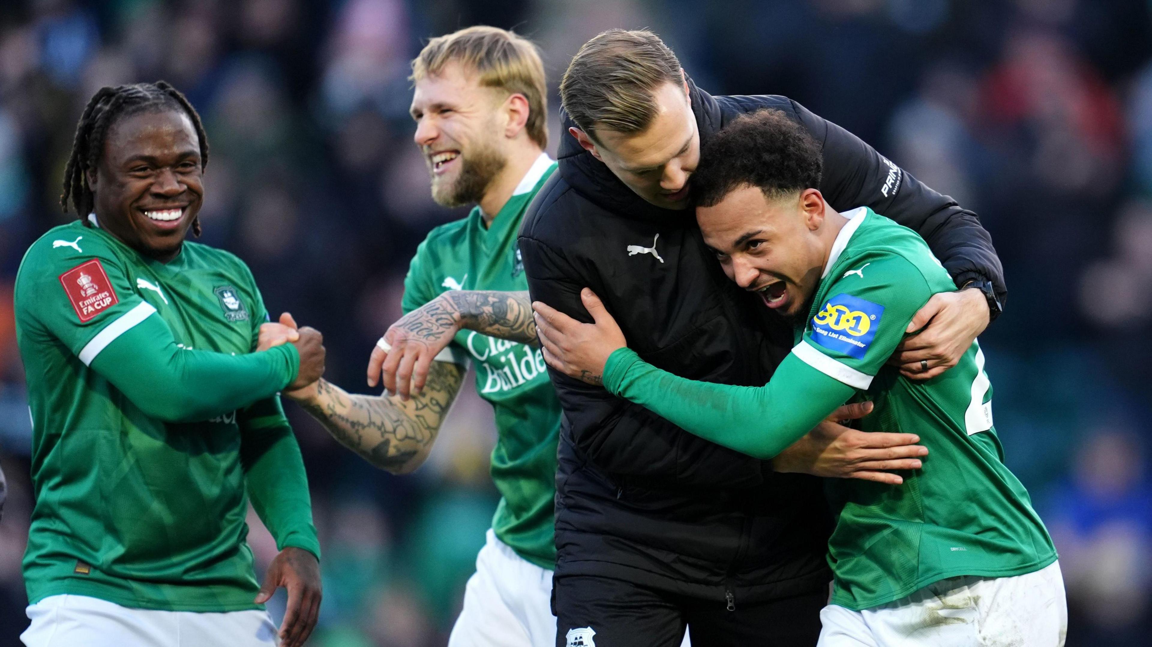 Four Plymouth Argyle players celebrate their victory over Liverpool in the FA Cup fourth round. Three of the players are in their kit while the other is wearing a tracksuit. Two players are hugging each other and the other pair are shaking hands.