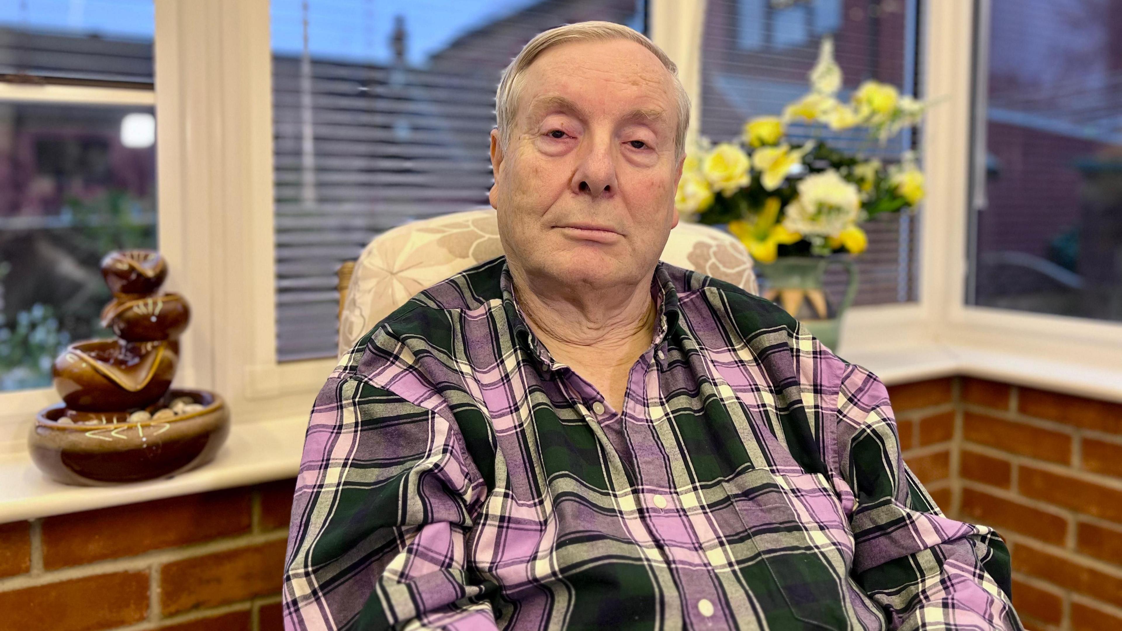 Mike Sarre, wearing a navy and purple checked shirt, looking into the camera from his flat in Norwich, with the window behind him