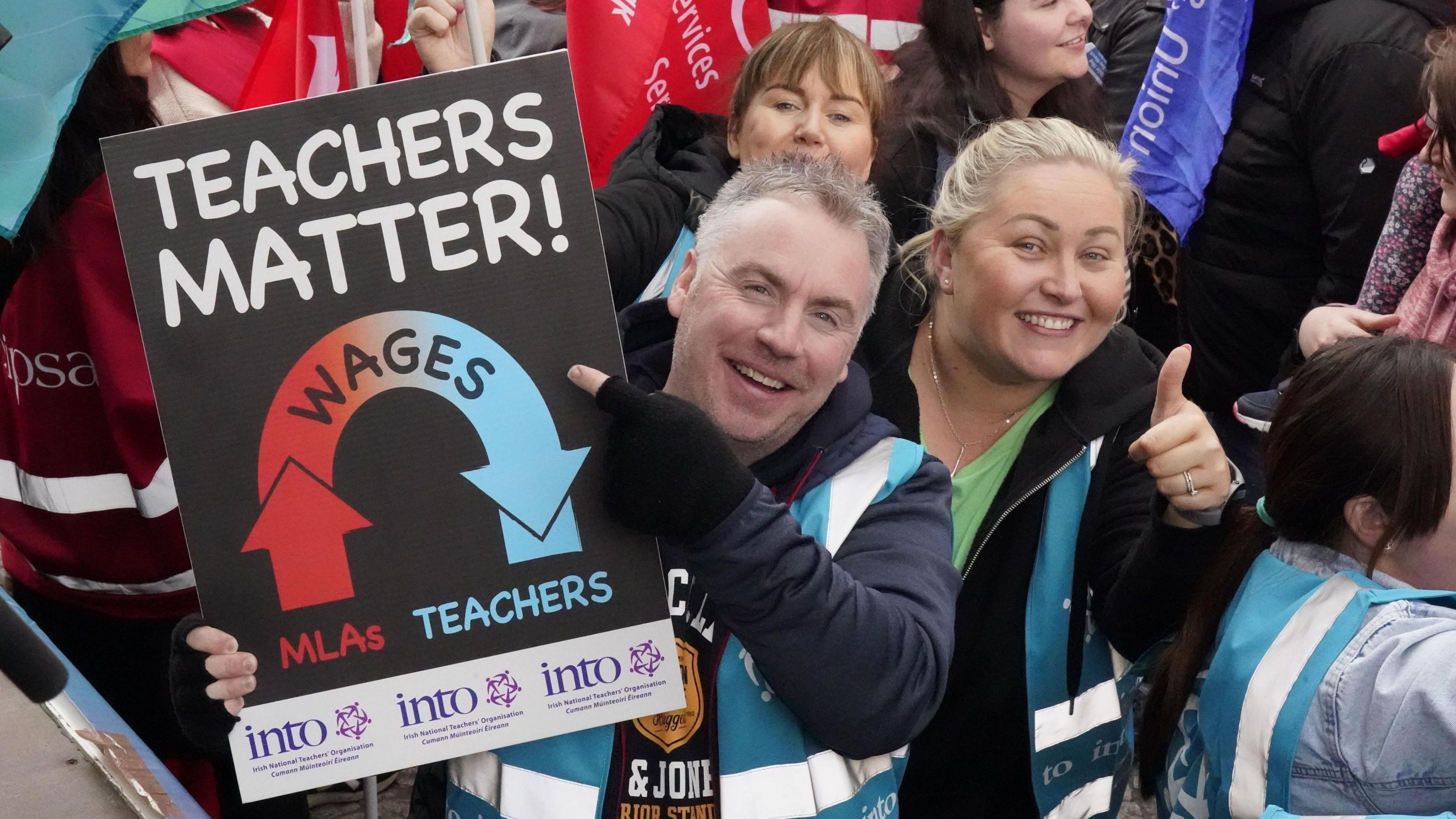 A man holding a sign that's reads "teachers matter!" and has an arrow with the word "wages" on it pointing from "MLAs" to "teachers" The Sign also has INTO branding. Beside him is a woman smiling and giving a thumbs up to the camera. They are standing in a crowd of people.