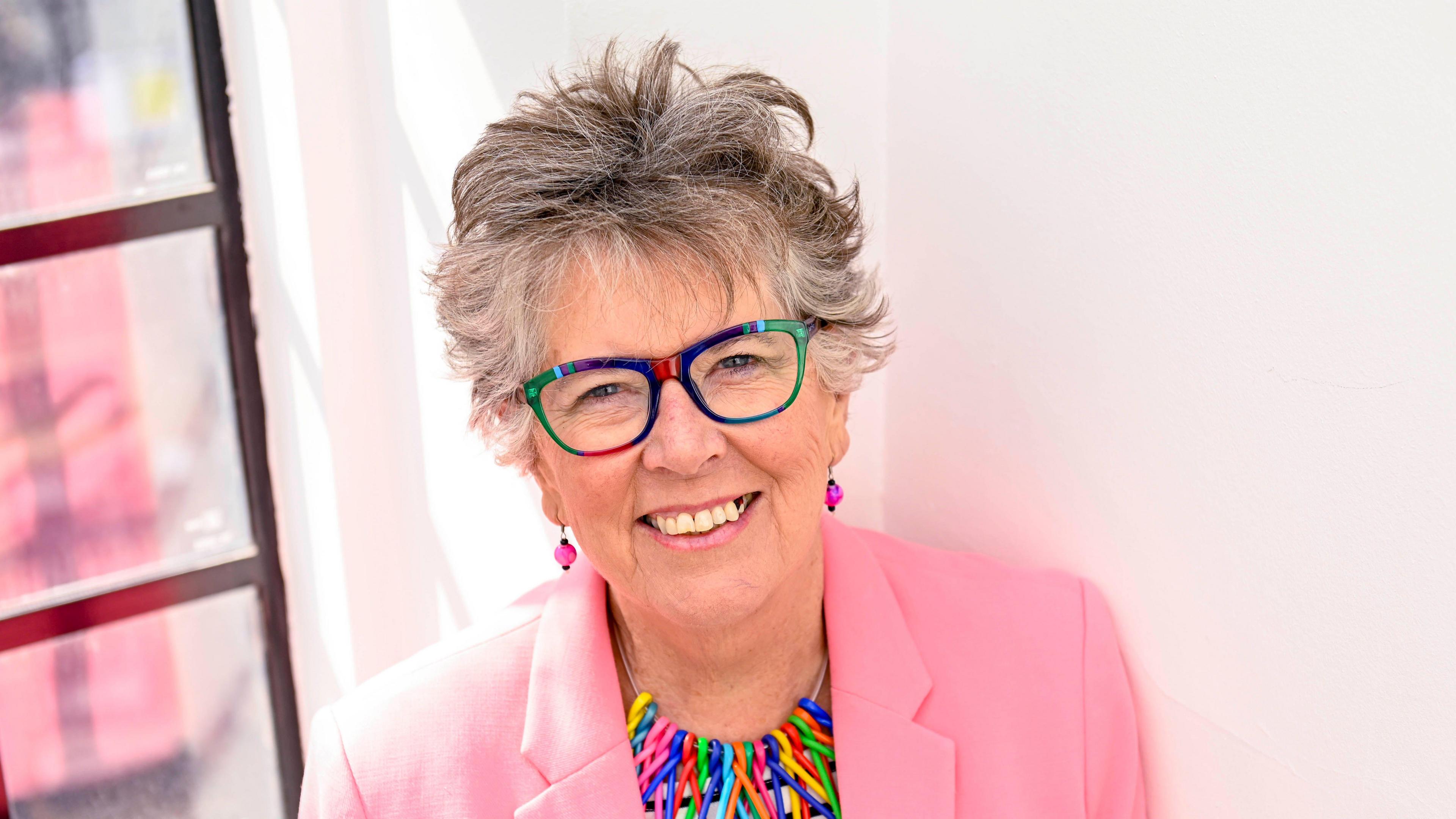 Prue Leith looks up into the camera, smiling against a bright background and wearing a pink blazer, pink earring and mulitcoloured glasses and necklace