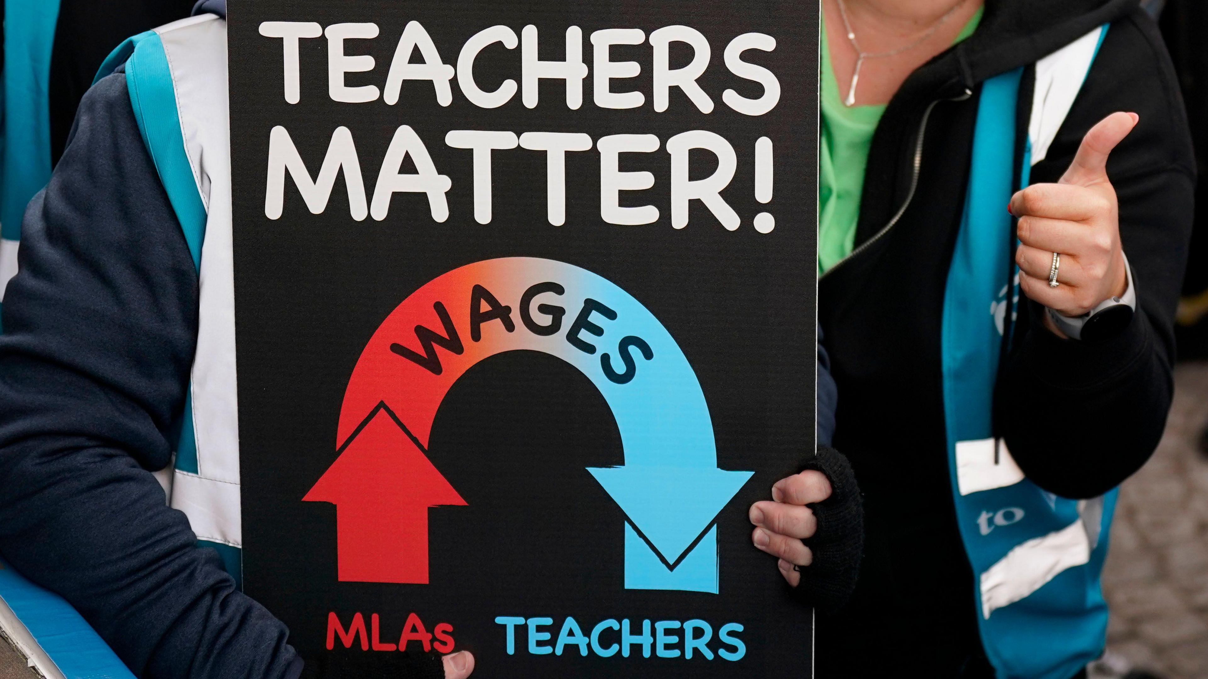 A placard saying 'Teachers  Matter', an arrow on one side is going up showing MLA wages and the other side is going down showing teacher wages.